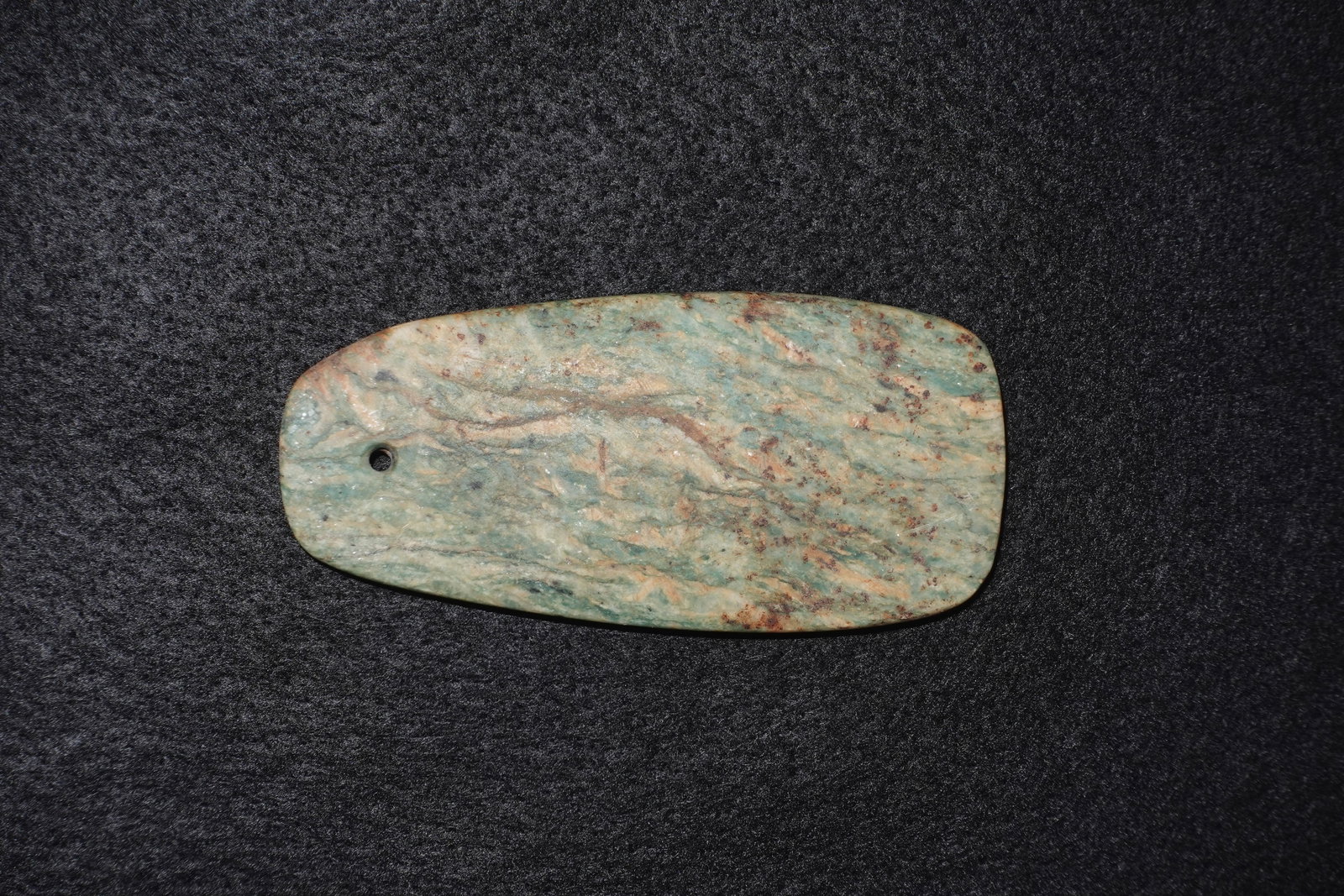 Chlorite Pendant: Green-blue chlorite carved into a flattened oblong pendant with soft rounded edges and a drilled suspension hole near one end. Surface displays marbled swirls of green, beige and brown with scattered