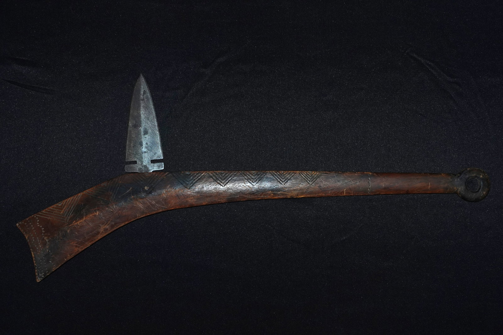 Plains Gunstock War Club: Gunstock-style war club carved from hardwood with a rounded ring handle at the butt. Repeated incised chevron and triangular punch motifs decorate the stock, which shows a dark patina and surface scra