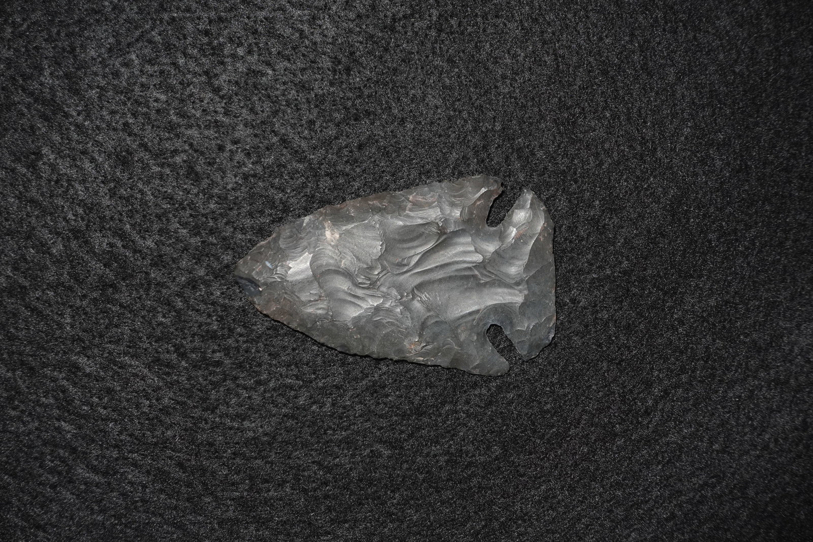 Thebes: Notched bifacial stone projectile point with a dark gray to black glassy surface and pronounced conchoidal flake scars. Broad triangular blade narrows to a notched base with deep lateral notches and