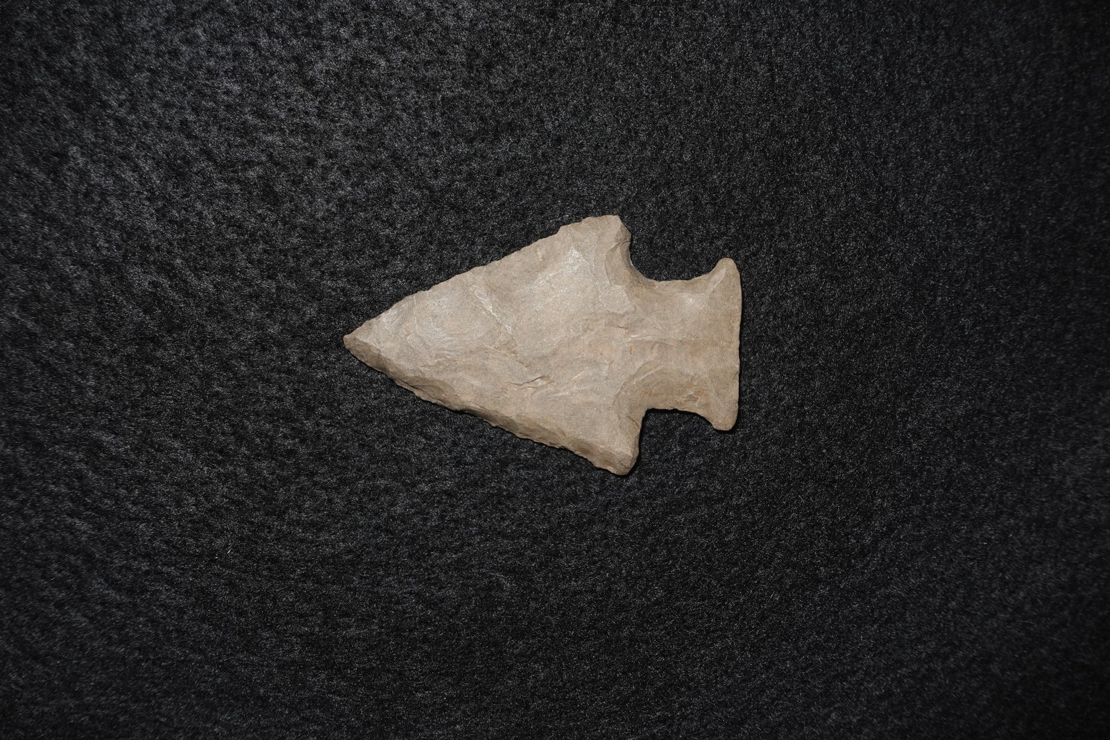 Stringtown: Flaked stone projectile point with a triangular blade, pronounced basal notches and broad shoulders. Light tan to beige coloration with extensive flake scars along the blade and a matte, fine-grained