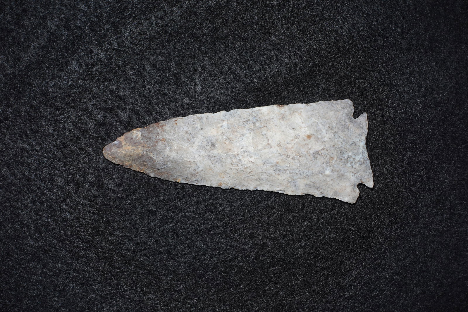 Decatur: Elongated triangular flaked stone projectile point with a long tapered blade and pronounced basal notches. Pale gray to off-white material displays brown iron staining near the tip and extensive