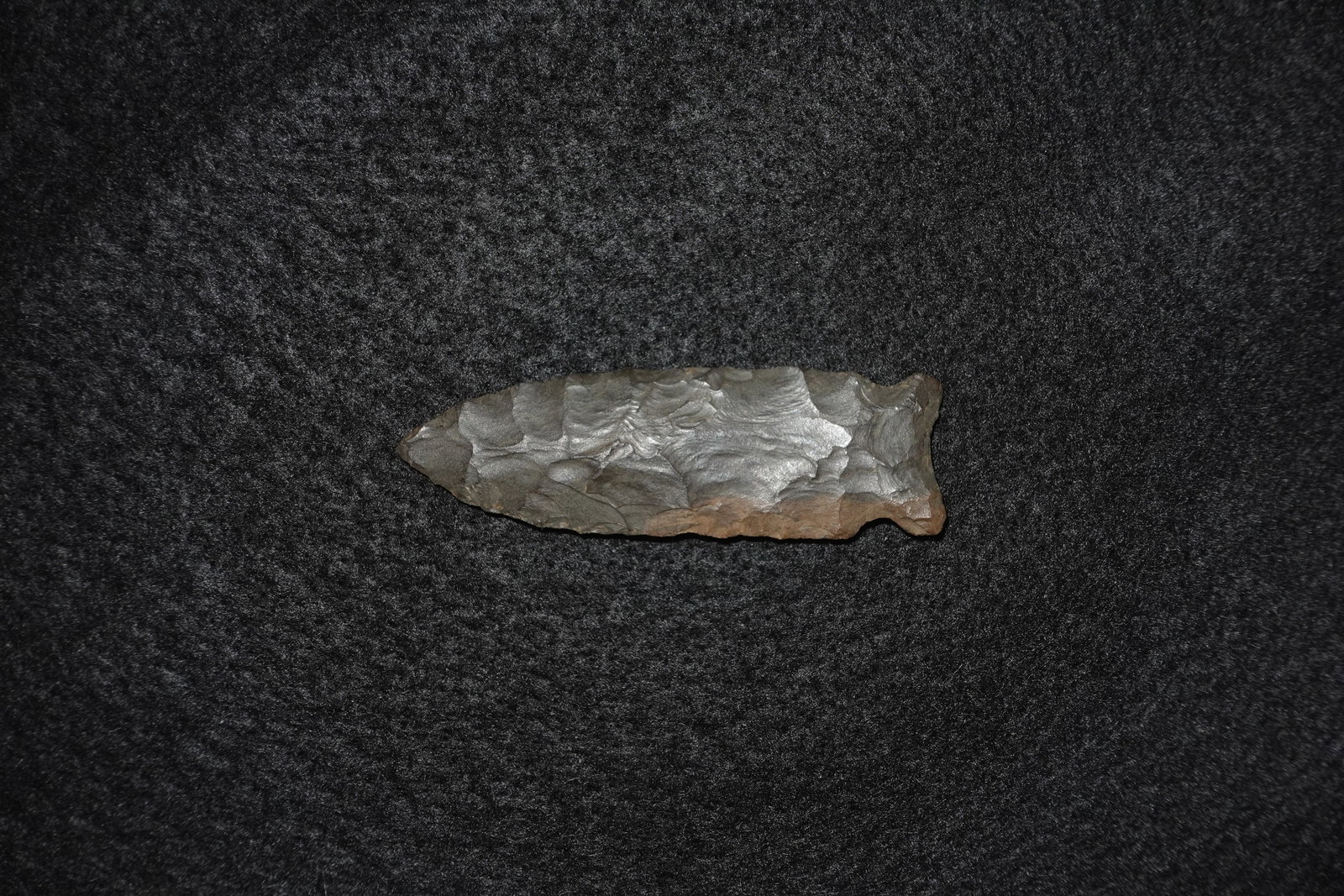 Benton: Elongated bifacial flaked stone point with pronounced faceted flake scars creating a leaf-shaped blade. Surface shows dark gray coloration with brown patina along the basal edge and clear