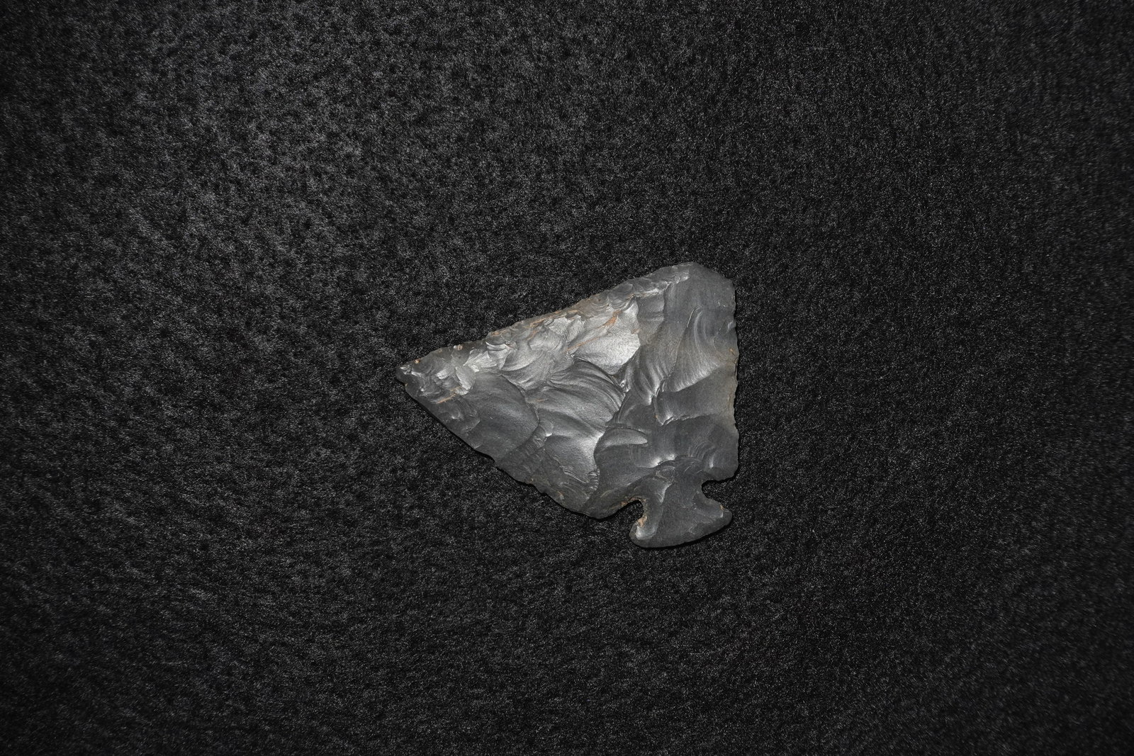 Corner Tang Knife: Corner-tanged knapped stone knife fashioned from dark gray to black knappable stone with pronounced conchoidal flake scars across both faces. Triangular blade narrows to a small basal corner tang