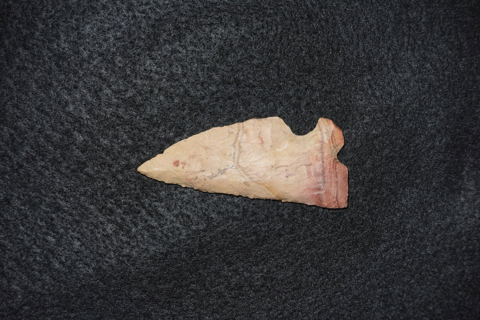 Corner Tang Knife: Corner-tanged stone blade with an elongated triangular profile and a pronounced basal corner notch. Bifacially flaked across a pale pink-to-tan stone with reddish banding, showing distinct flake