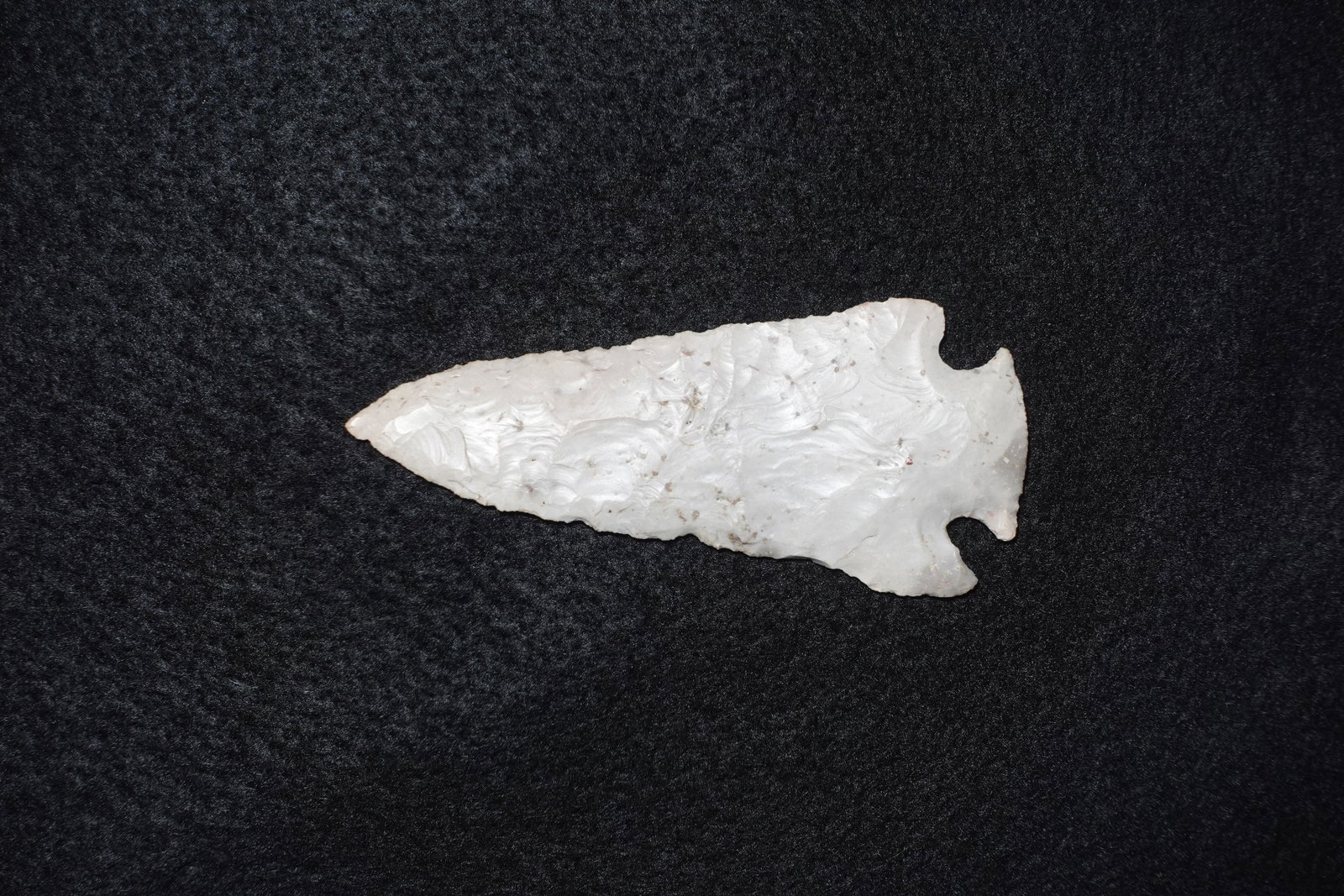 Kay Blade: Long white flaked stone blade with a lanceolate triangular outline and a notched stem base. Bifacial flaking produces overlapping ripple scars across creamy translucent material with small mineral