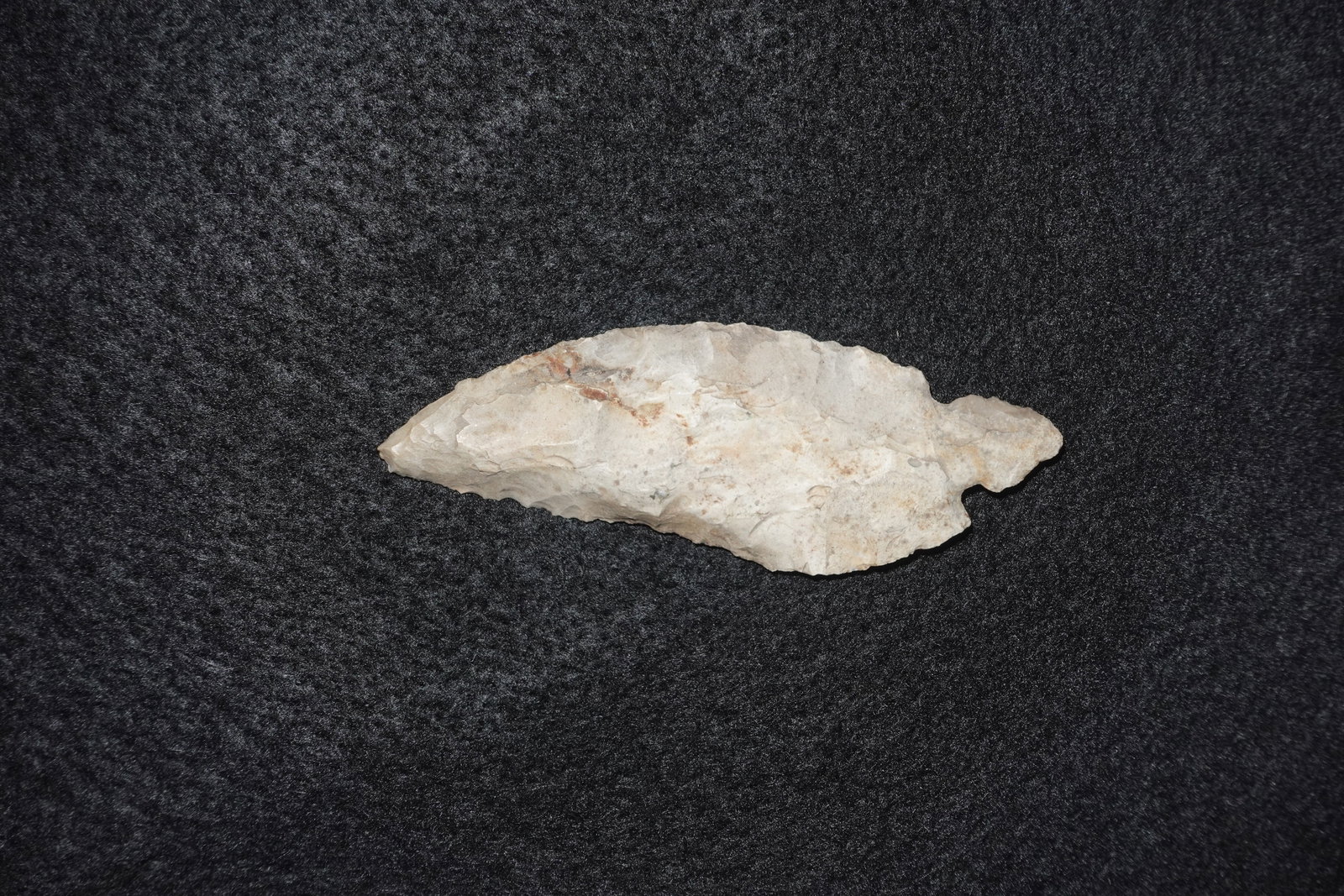 Turkeytail: Leaf-shaped flaked chert point with an elongated, slightly recurved profile and a tapered tip. Light cream to tan coloration with subtle gray and brown staining across the surface, showing bifacial fl