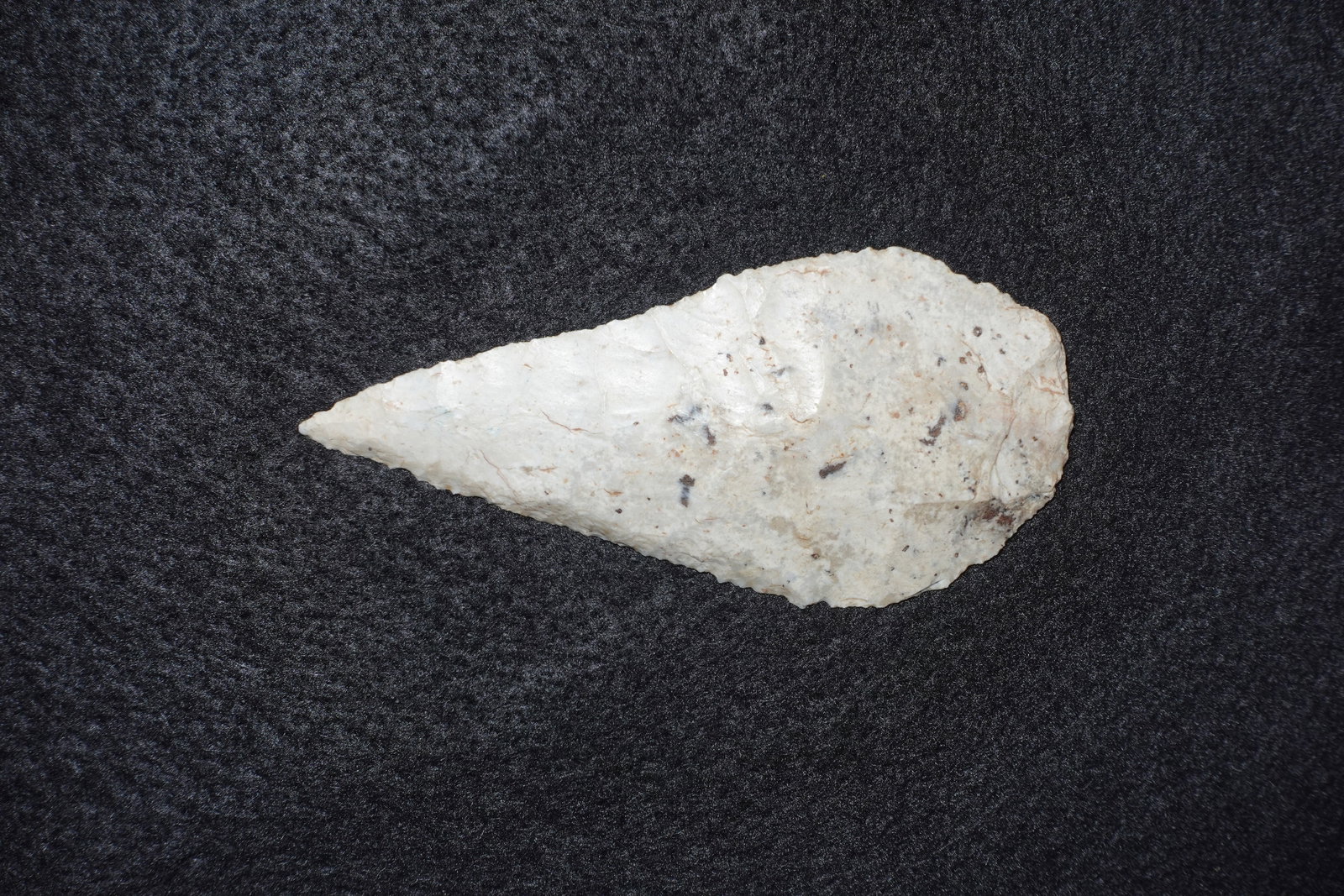 Cobbs Blade: Elongated triangular stone blade in a cream-to-white hue with scattered dark mineral specks throughout the surface. Bifacially flaked with fine pressure-flaked edge serrations, a sharply pointed tip a