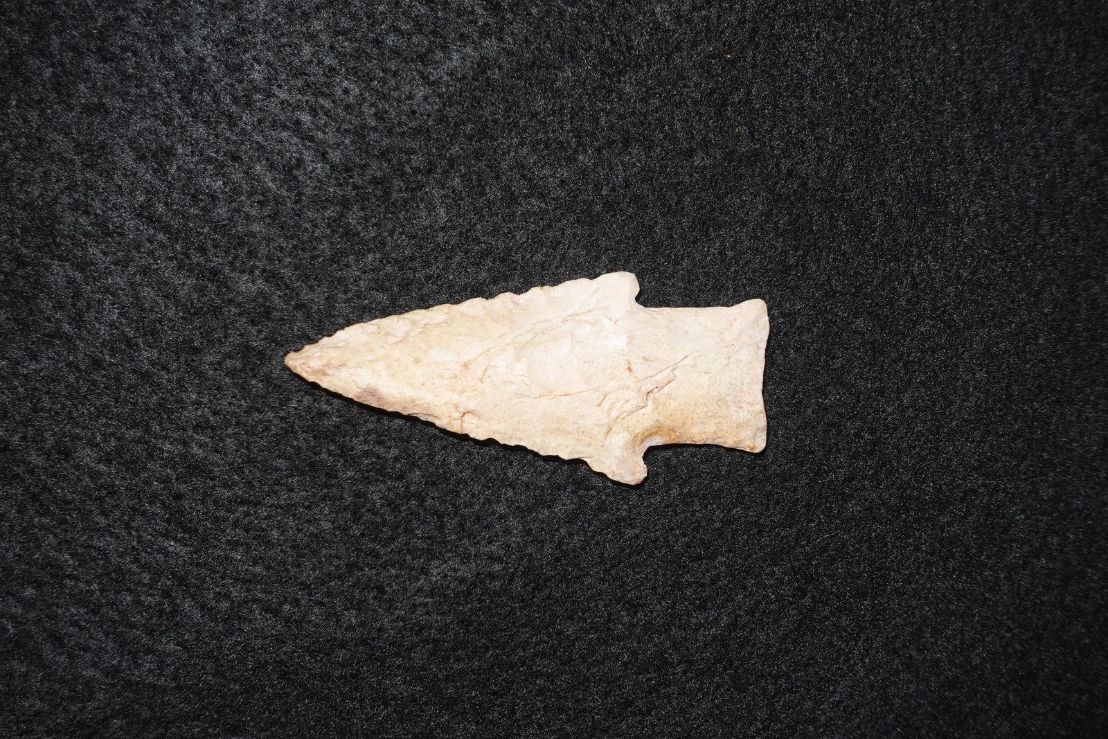Hardin: Light tan-to-cream stone projectile point with bifacial flaking and serrated marginal edge. Triangular blade tapers to a sharp tip and broadens to a stemmed, notched base with a small shoulder.