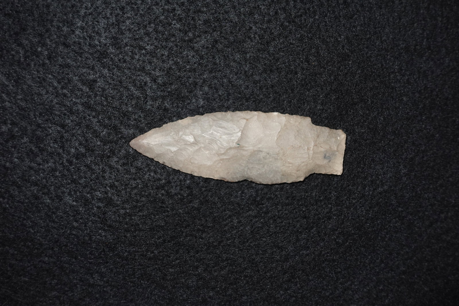Kramer: Long lanceolate bifacially flaked stone projectile point with a narrow stemmed base and worked tang. Light gray to tan coloration with visible flake scars and subtle mineral flecking. Blade exhibits