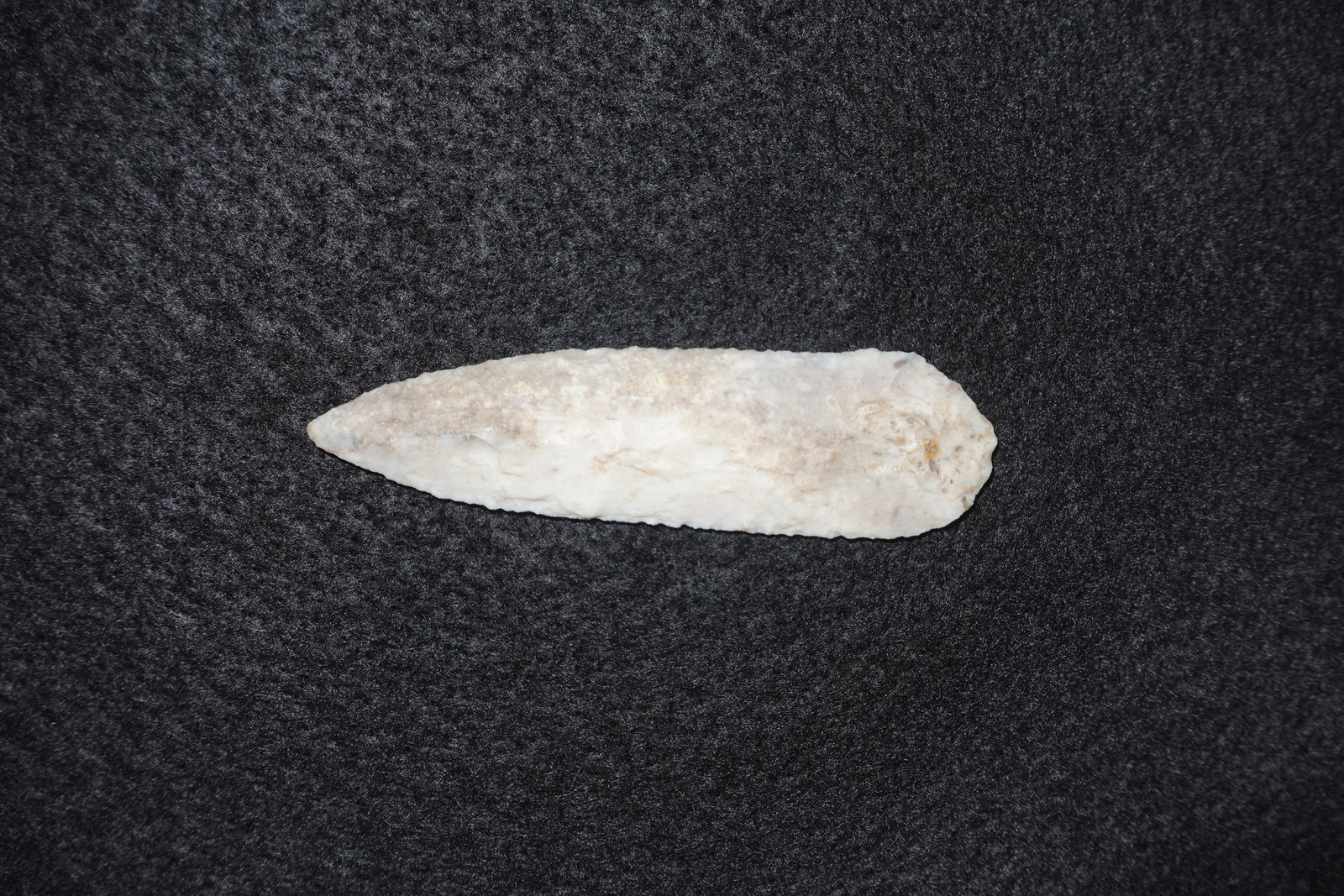Paleo Blade: Elongated, leaf-shaped flaked stone blade with a tapered point and rounded base. Bifacial flaking across both faces produces distinct flake scars and fine edge retouch, with pale white to cream colora