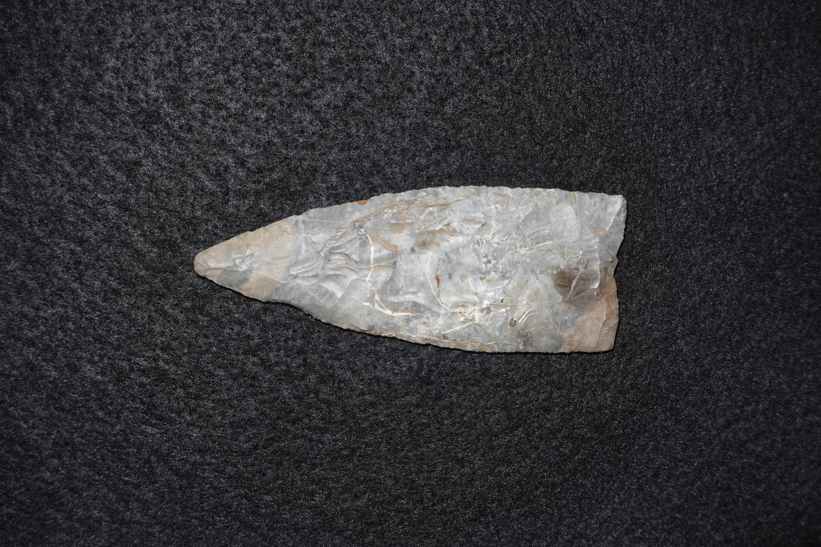 Clovis: Elongated bifacial flaked stone point with a lanceolate profile and a sharply tapered tip. Gray chert shows closely spaced pressure flake scars across both faces and light brown staining along the
