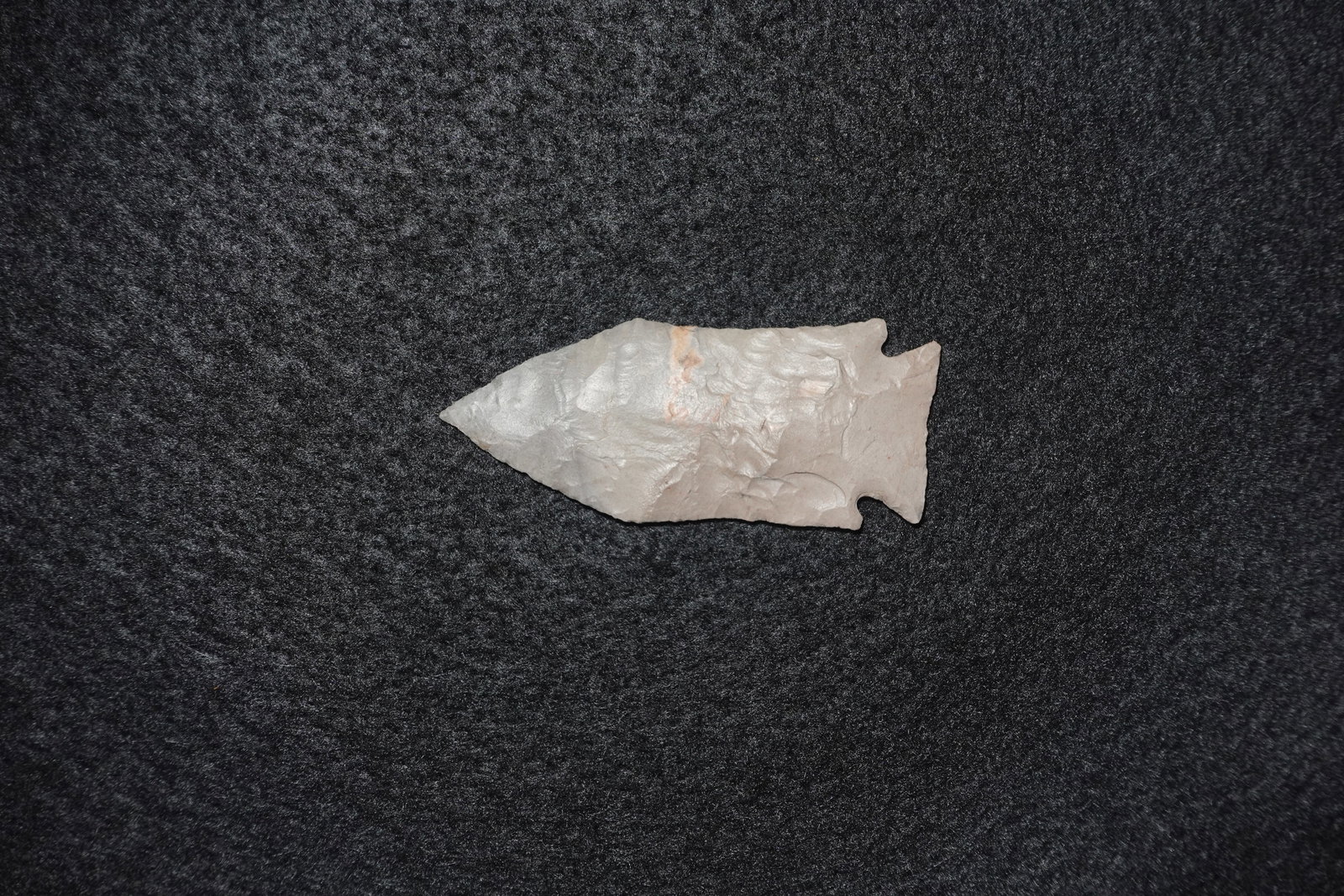 Afton: Bifacially flaked stone projectile point with a triangular blade and a notched, barbed base. Light gray to tan material with dense flake scars and fine pressure-flaked edge work, terminating in a