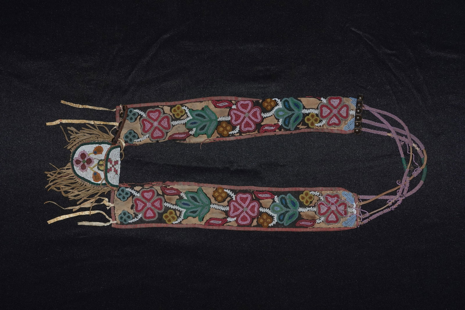 Early Cree Shot Pouch: Beaded Cree shoulder pouch featuring floral seed-bead panels along twin fabric straps and a fringed hide pouch. Floral motifs executed in pink, green, red, yellow and white glass seed beads are