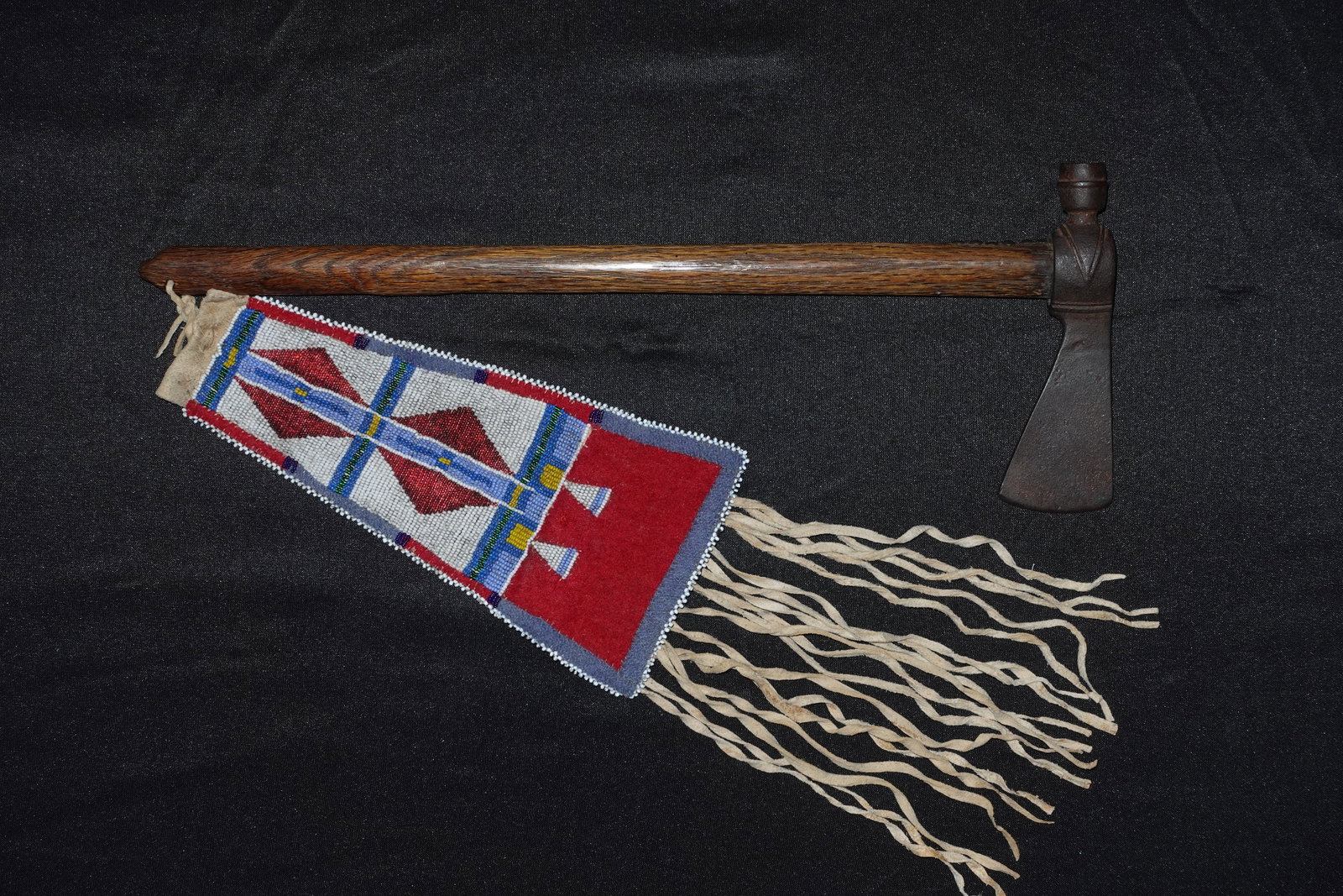Crow Pipe Tomahawk w Beaded Drop: Pipe tomahawk combining an iron axe head with an integrated pipe bowl at the poll, mounted on a straight stained wooden haft with carved knob terminals. Shaft includes a row of brass tacks and a fitte