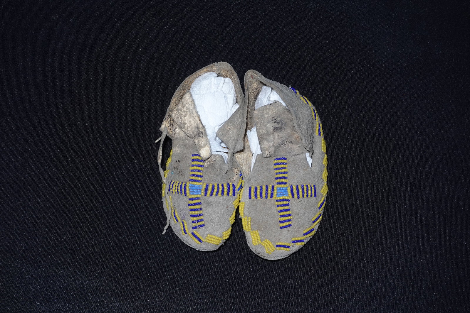 Southern Plains Moccasins: Hand-stitched soft leather moccasins decorated with yellow, dark blue and light blue glass beadwork arranged in cross/grid motifs across the vamp and along the edge. Natural gray-tan hide exhibits