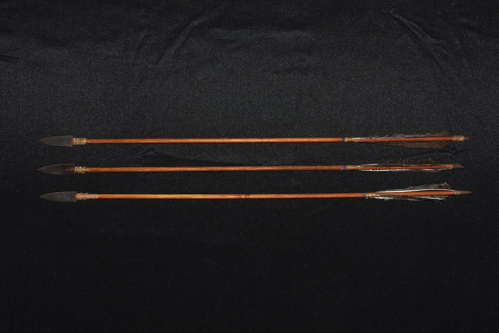 Set of 3 Primitive Arrows: Three primitive-style arrows featuring straight polished brown wooden shafts fitted with dark steel points. Dark brown feather fletching is secured with sinew or twine at the nocks, and sinew wraps