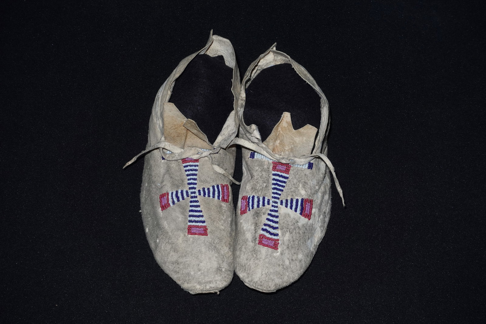 Southern Plains Moccasins: Plains-style moccasins crafted from soft tan leather with black felt insoles and cross motif beadwork in blue, white, and pink on the vamp. Leather ties thread through the uppers and the flat leather 