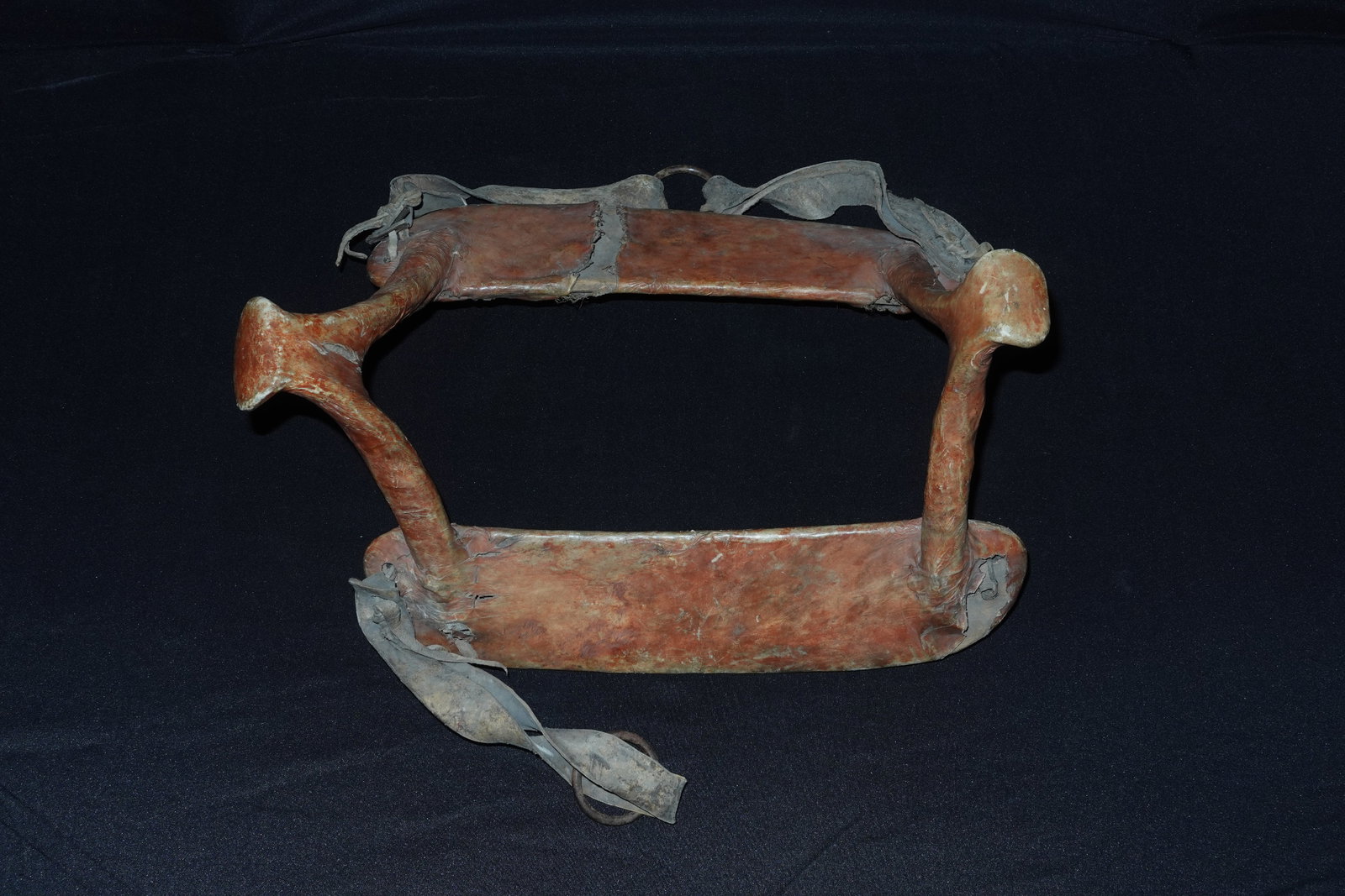 Northern Plains Saddle: Red ocher-toned leather stretched over a wooden saddle frame with paired antler supports at the pommel and cantle. Rawhide thongs and a metal O-ring secure the rigging, and seams are hand-stitched