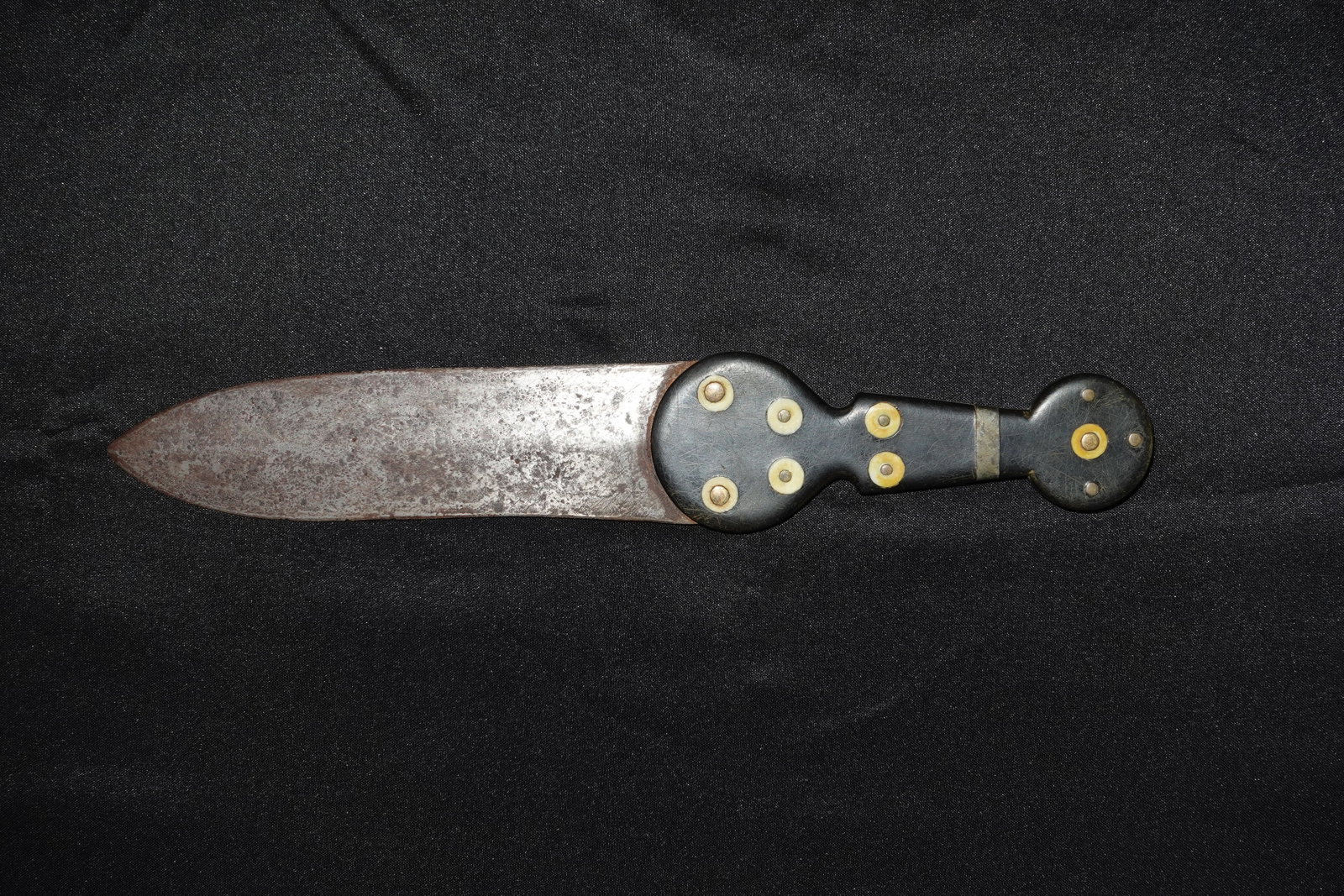 Dag Knife: Broad single-edged steel blade with pronounced surface pitting and a gray-brown patina across the cutting surface. Black wooden handle with decorative circular bone inlays set into the scales and