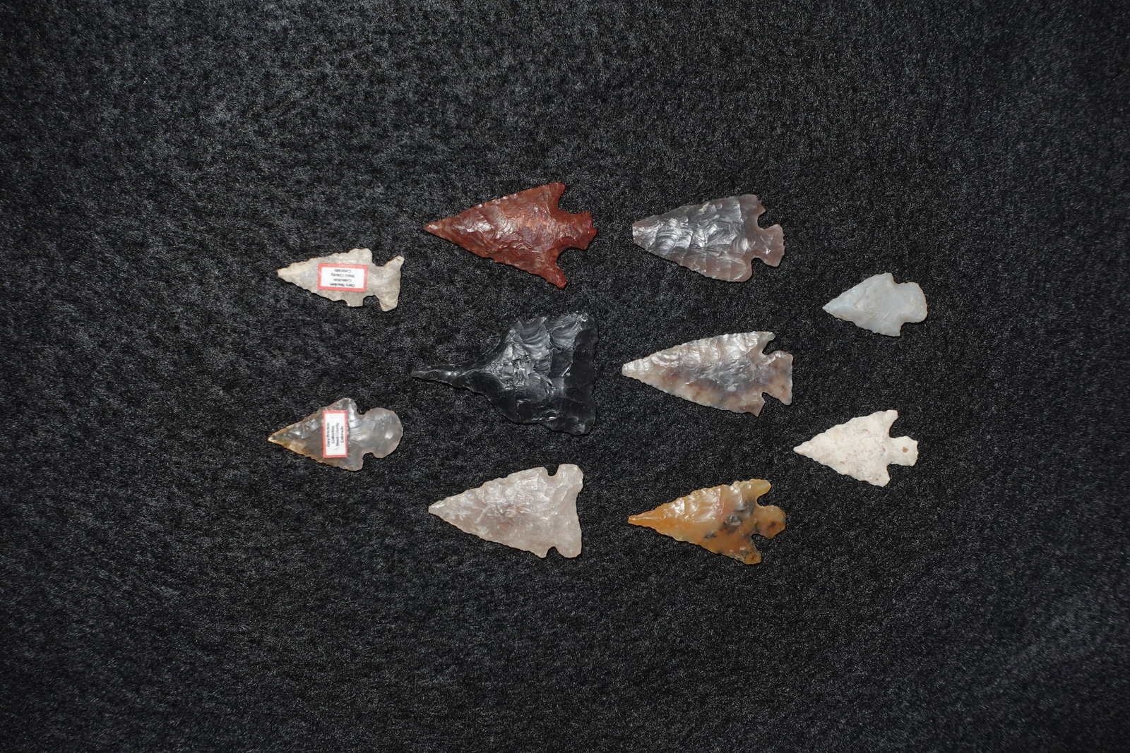Group of Western and High Plains Gem Points: Assortment of gem points fashioned from translucent to opaque flaked stone, featuring a range of colors from deep black and rust red to pale gray and amber. Pieces exhibit fine bifacial flaking, visib