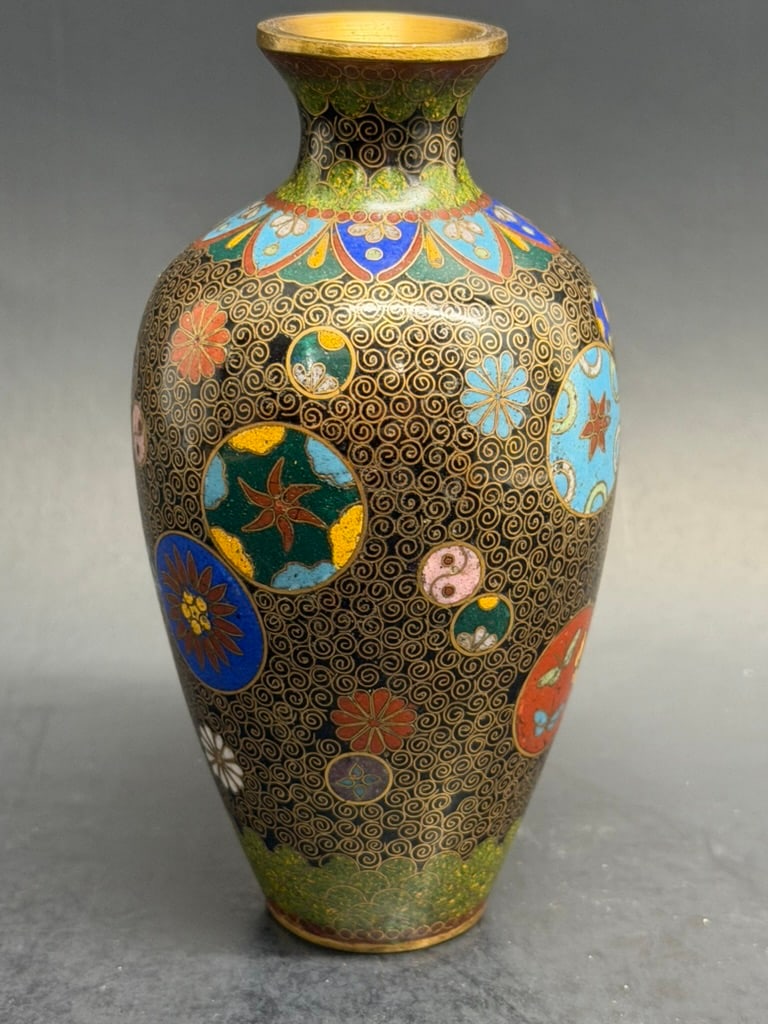 Japanese Meiji Period Cloisonne Vase unsigned: A small slightly lobed cloisonne enamel vase with roundels containing flowers and Geometric shapes amist spiral wire over black ground. Some highlights in Goldstone. Gilt wires, rim and base. Meiji Pe