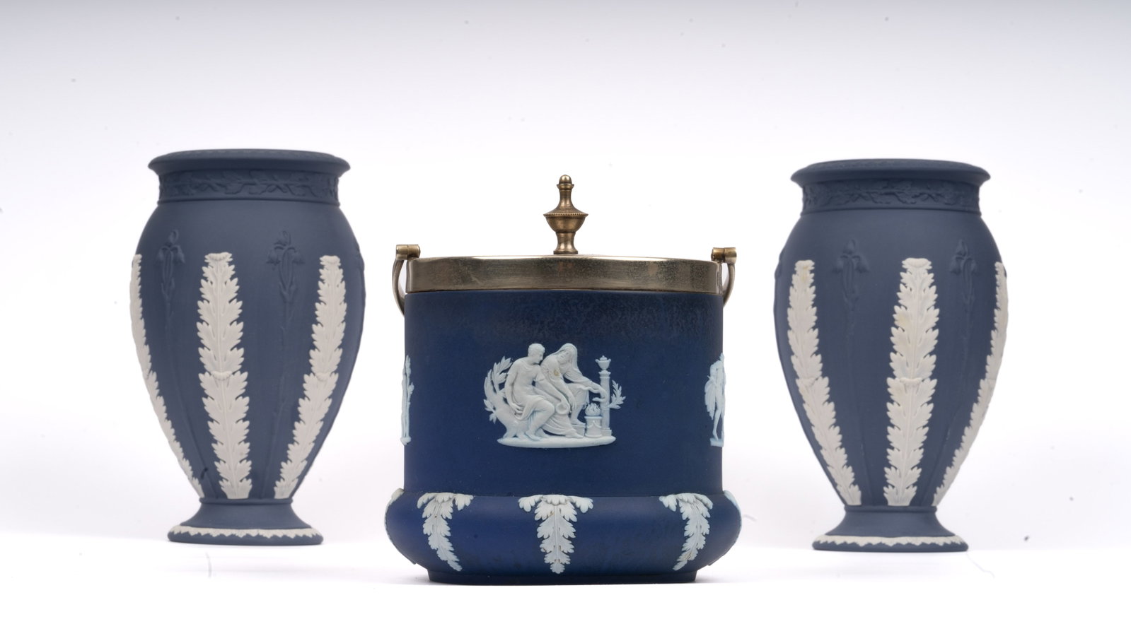 Pair Wedgwood Jasperware Vases and Biscuit Jar: Comprising a pair of classical Wedgwood jasperware vases with applied white foliate reliefs on matte cobalt ground, and a matching biscuit jar with applied figural scenes in neoclassical style. The bi