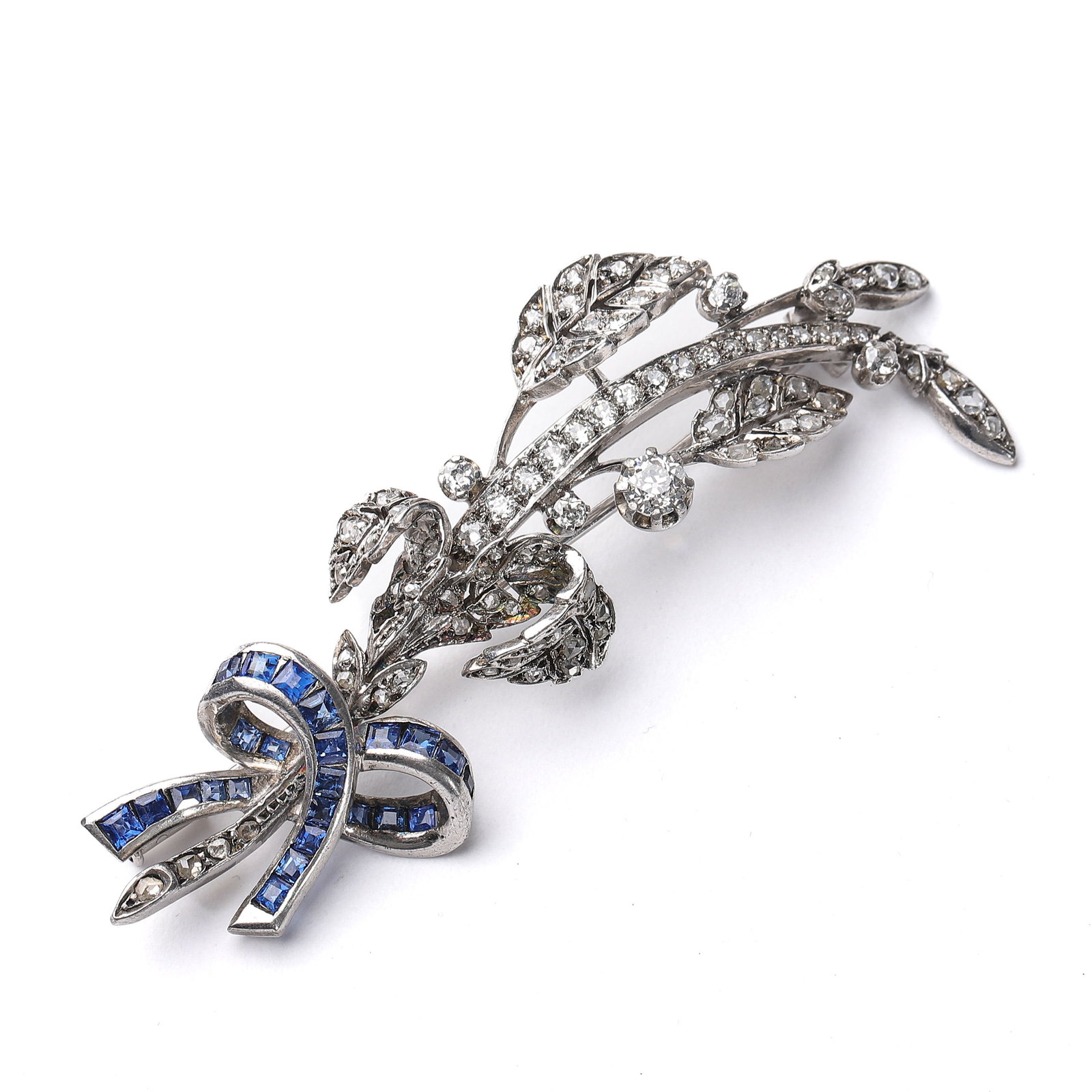 ART DECO FRENCH GOLD AND SILVER-TOPPED DIAMOND BROOCH: An exceptional Art Deco (1925-1935) brooch, meticulously handcrafted in 14K white gold with silver overlay. This elegant piece features a seamlessly channel-set blue sapphire ribbon bow, creating a