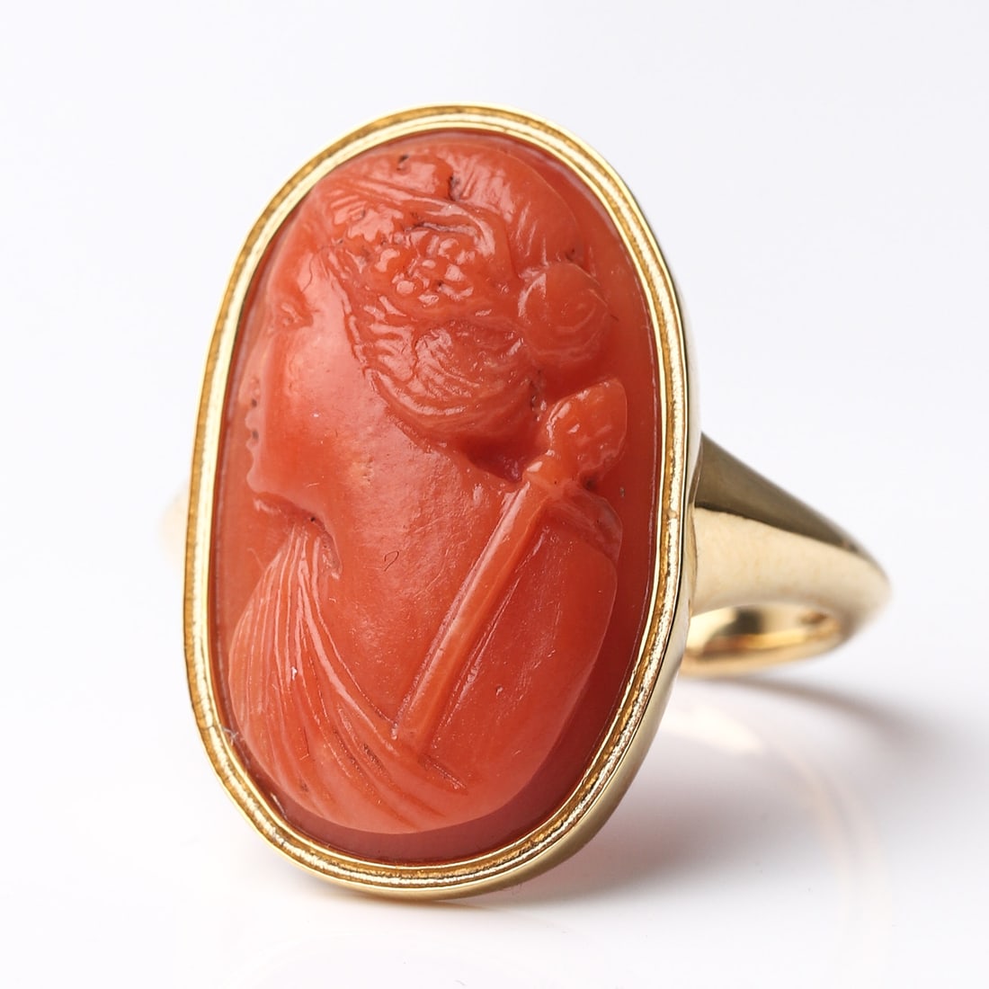 A GOLD RING WITH CORAL CAMEO: Dimensions Inner diameter 18.7 mm; face 1 × 5/8 in (24.5 × 15.8 mm); weight approx. 6.8 g. Description: The oval cabochon coral, carved in relief with the bust of a classical maiden in profile, her
