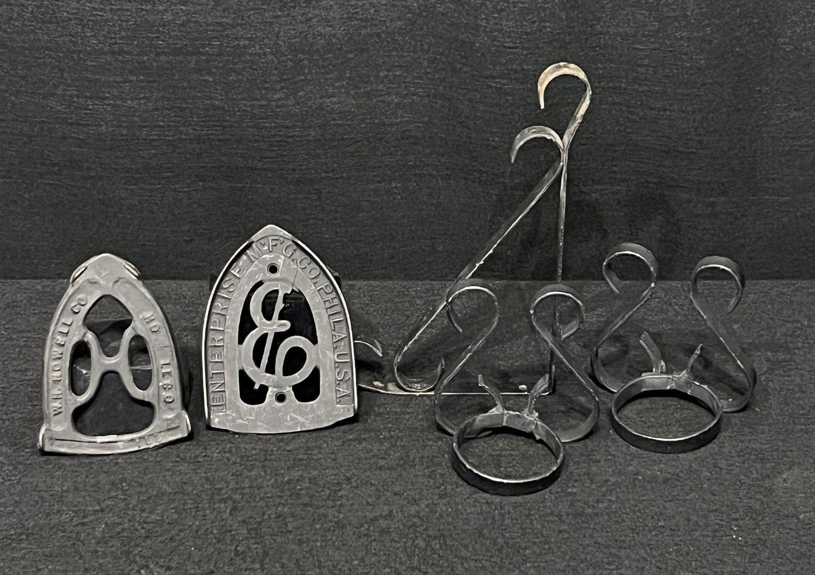 Cast Iron Sad Iron Trivets, Metal Plant Hanger & Wrought Iron Decorative Holders: These cast iron sad iron trivets are inscribed with the manufacturing names, Enterprise Mfg and W.H. Howell Co. One trivet tray shows a crack near the top. It is still stable but the crack is visible.