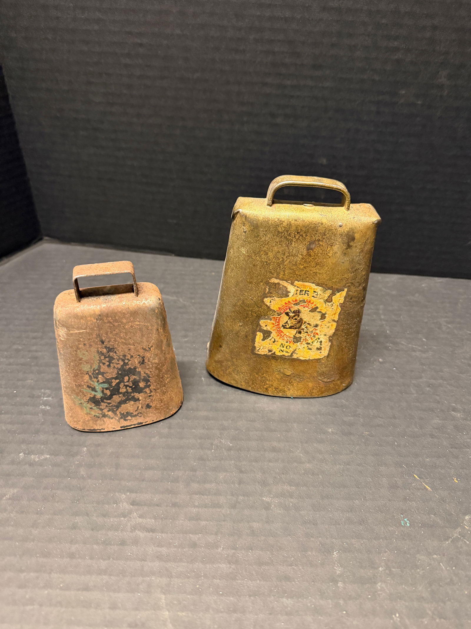 Antique livestock / cow bells (2): Antique livestock / cow bells (2), one larger with original partial paper label, heavy riveted iron construction with top handles, likely late 19th–early 20th c.; rustic farm patina throughout.