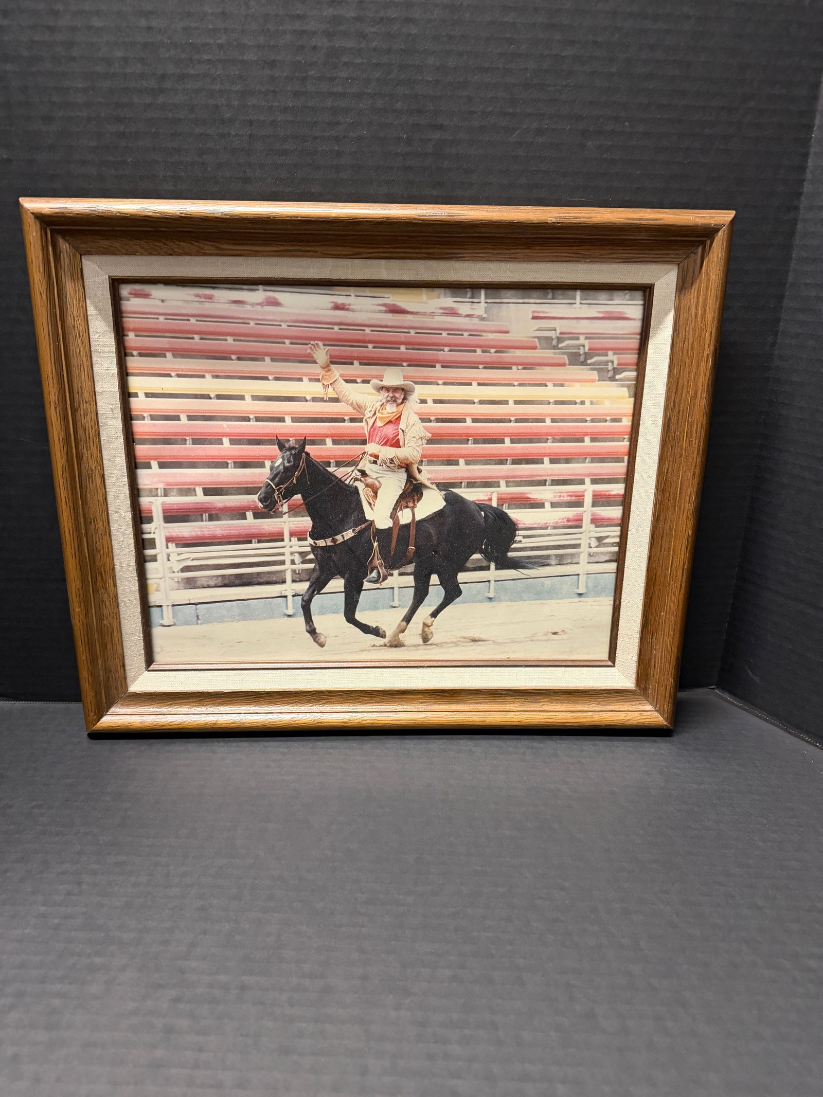 Framed Western cowboy photographic print: Framed Western cowboy photographic print depicting bearded rider on black horse waving in rodeo arena, wood frame with linen liner, color photograph, approx. mid-to-late 20th century, overall 14&rdquo