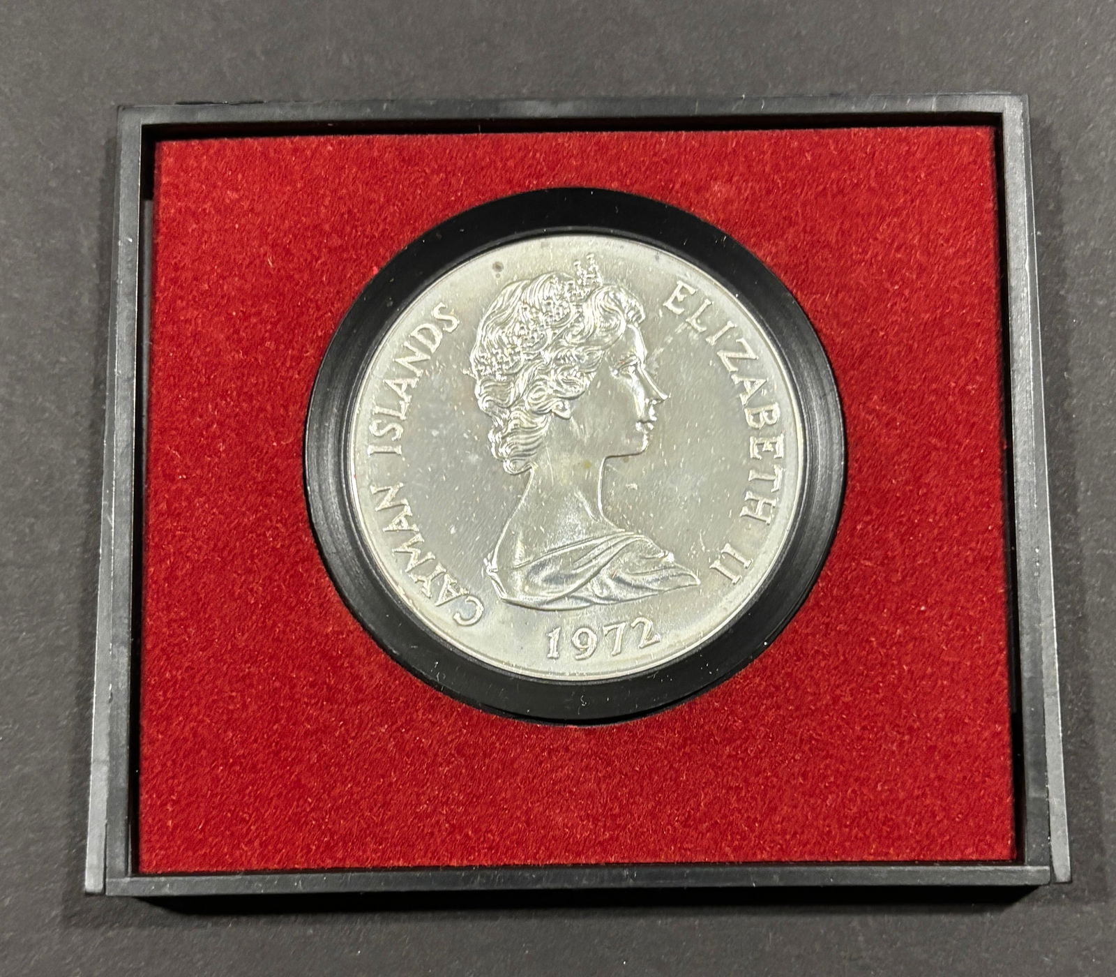 1972 Cayman Islands Crown Size Commemorative Coin: 1972 Cayman Islands commemorative crown-size coin depicting Queen Elizabeth II, presented in fitted red velvet display holder. Reflective surfaces with scattered light handling marks and mild toning