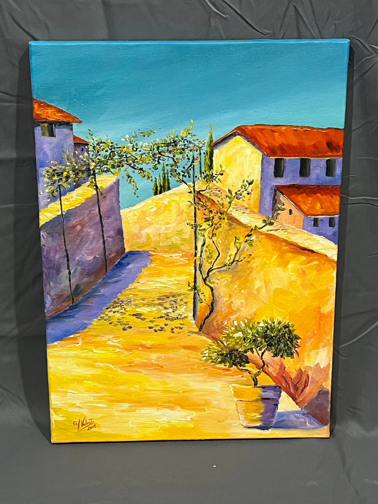 46Signed Manuela Valenti Village Scene Oil on Canvas: This image shows a contemporary acrylic or oil painting depicting a sun-drenched Mediterranean village street.Artistic AnalysisSubject Matter: A narrow European village lane, characteristic of