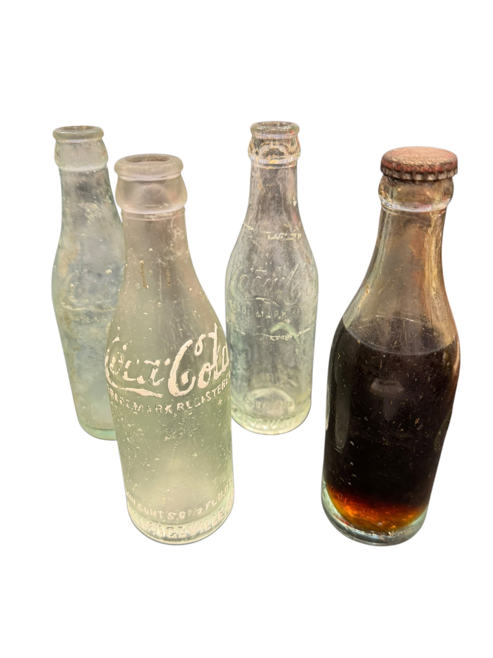 Lot of 4 Straight Side Coca Cola Bottles: The image shows a collection of vintage clear and green-tinted glass Coca-Cola bottles, including three weathered or empty bottles and one intact bottle filled with dark liquid and a rusted crown