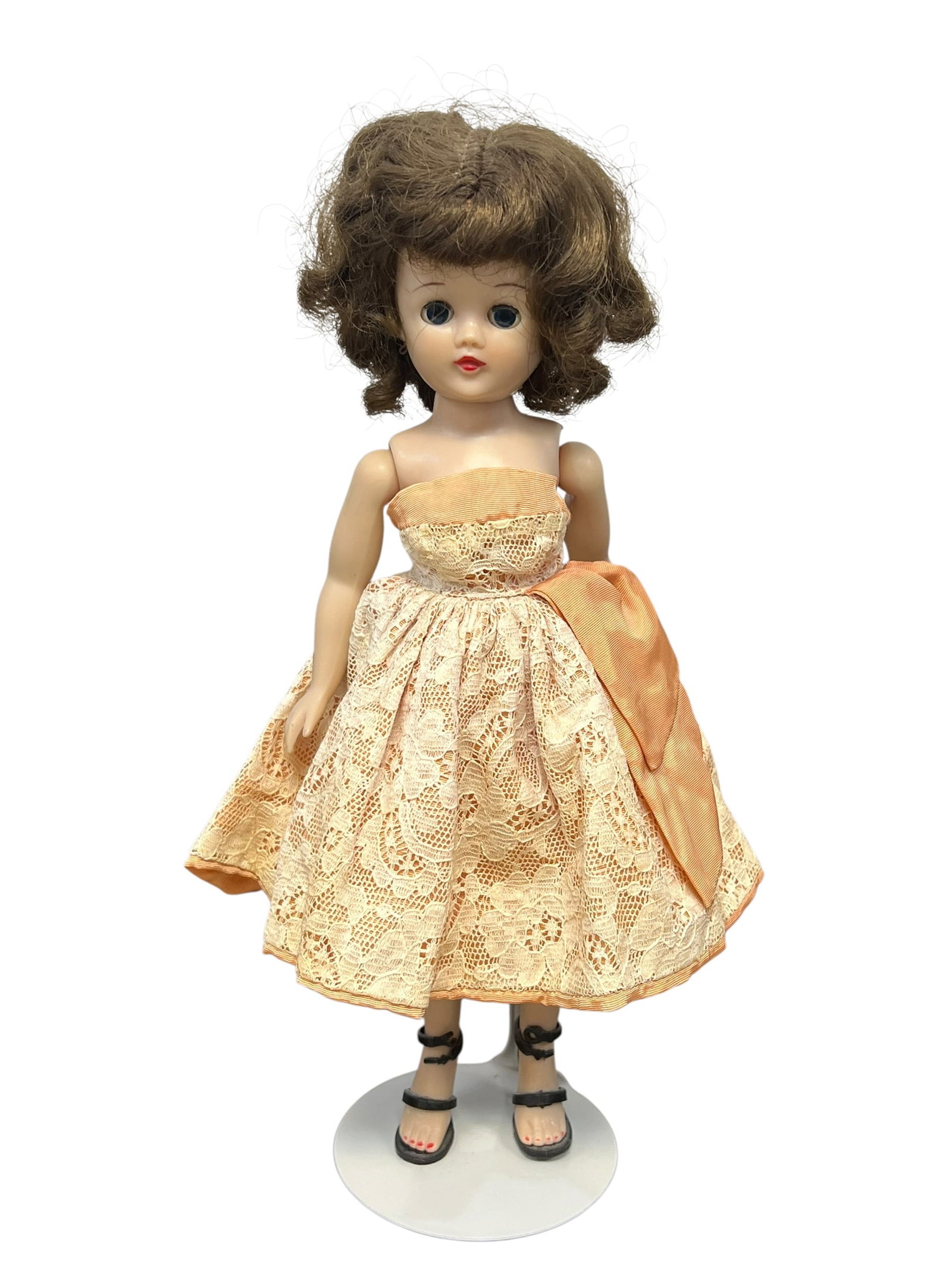 Vintage Vogue “Jill?? Plastic Doll: This image shows a vintage 1950s fashion doll, likely a Vogue Jill or an Ideal Miss Revlon doll, wearing an peach-colored lace dress with a matching side bow and black high-heeled sandals. [1, 2, 3]Do