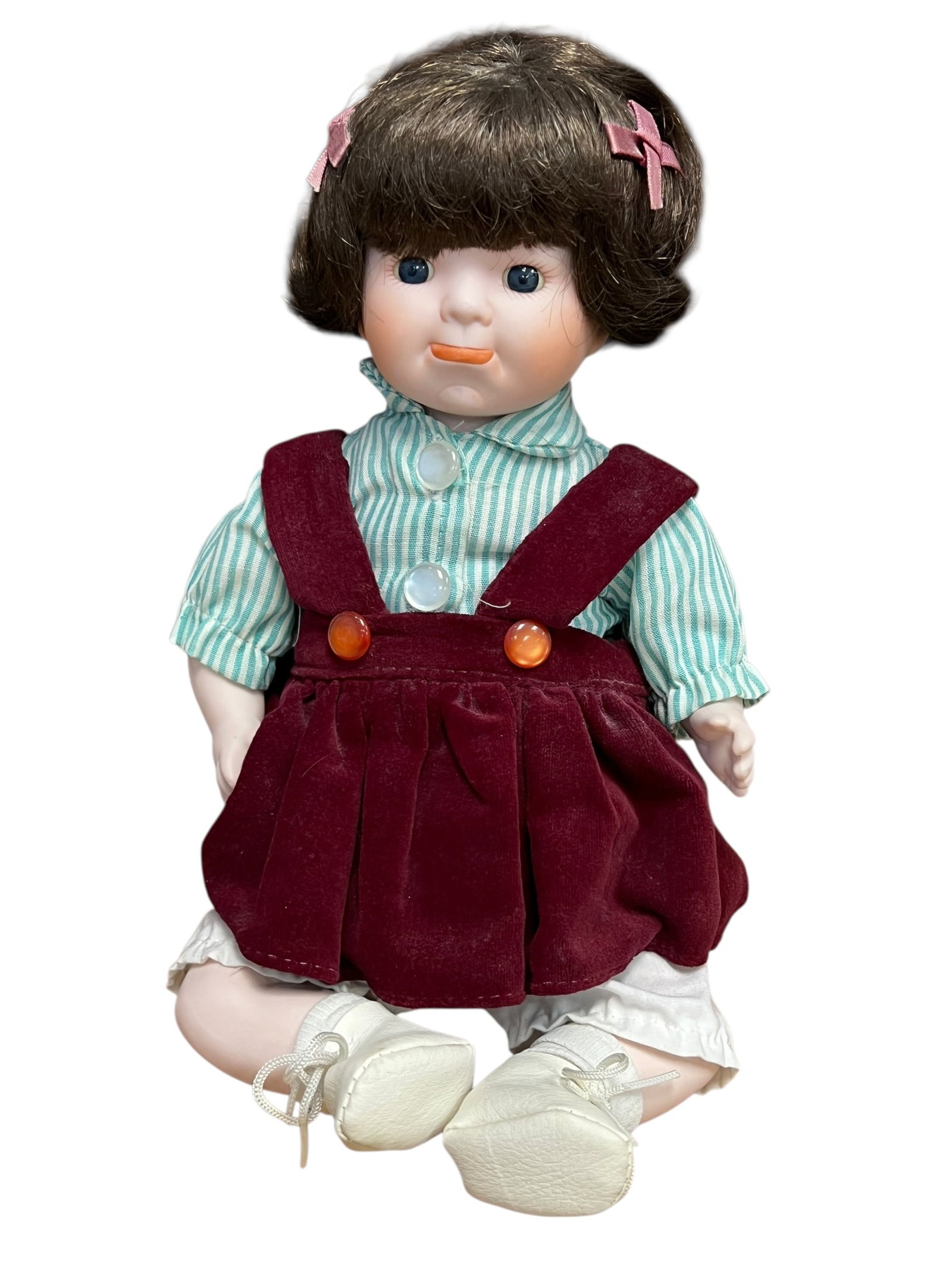 Bisque Head Doll: The image shows a vintage collectible porcelain doll with brown bobbed hair, pink hair clips, blue eyes, and a green-and-white striped shirt under a burgundy corduroy overall dress.Because