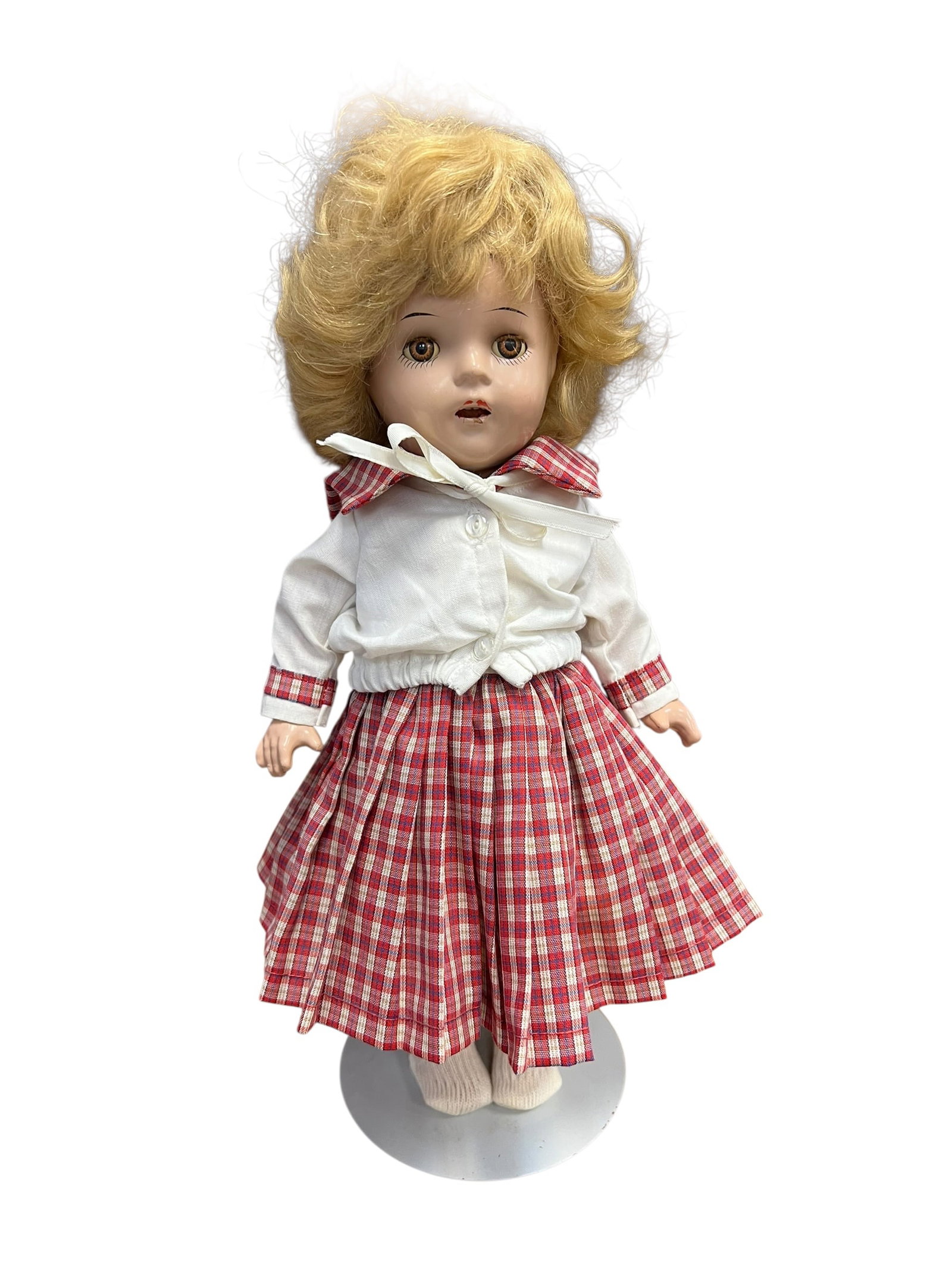 Composition Doll: vintage porcelain collector doll with short curly blonde hair, brown sleeping eyes, and an open-mouth expression, wearing a white cardigan and a red-and-white plaid skirt on a metal display