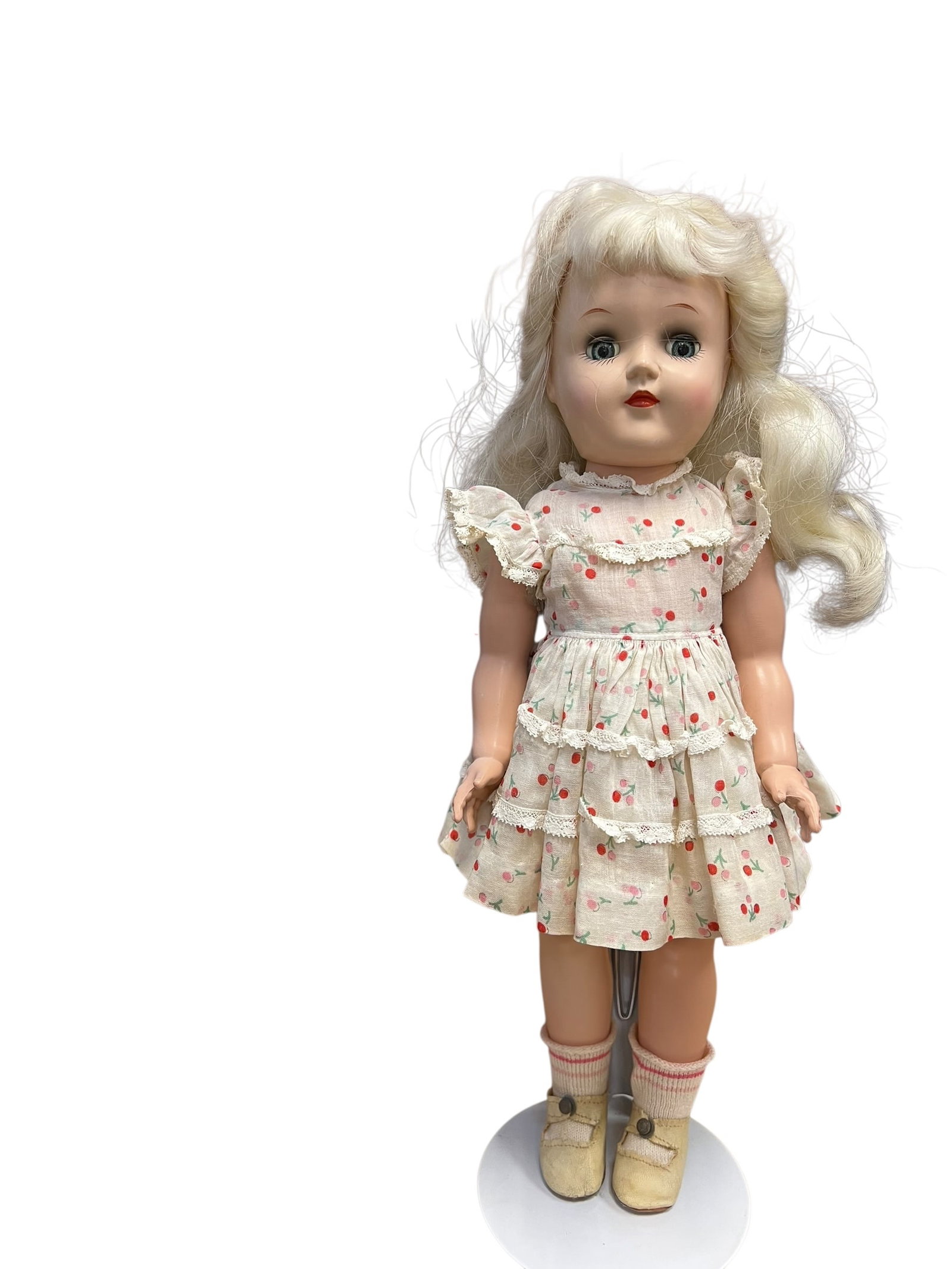Ideal Plastic Doll: vintage 1950s Ideal Toni Doll with blonde hair, sleep eyes, and a floral-patterned ruffled dress. [1, 2, 3]Type: Hard plastic collectible doll with jointed limbs.Origin: Introduced by the Ideal