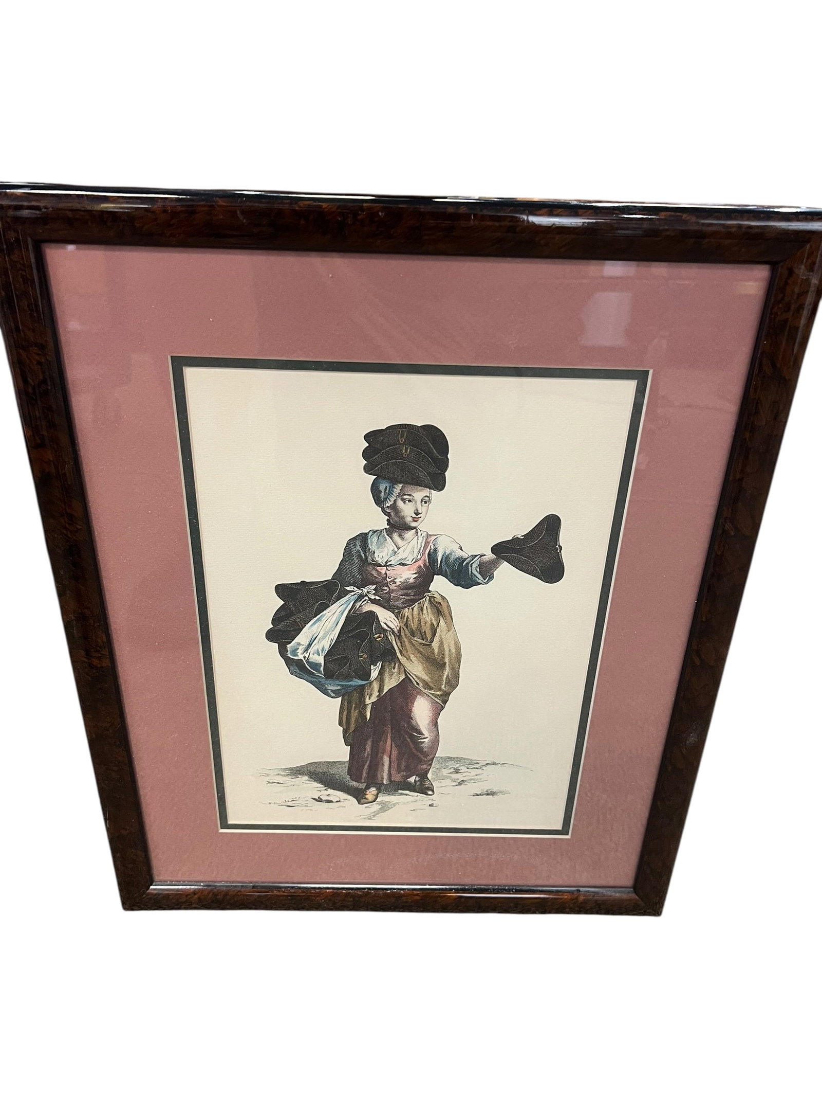 Framed Hand-Colored French Shoe Seller / Marchande de Souliers Print: Decorative framed French-style hand-colored print depicting a female shoe seller or street vendor, shown carrying an assortment of shoes while presenting one in her outstretched hand. The woman is dre