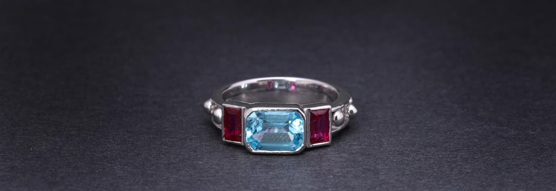 18k White Gold Ring by Carlo Rici: An 18k white gold designer ring by Carlo Rici, from a limited 1990s design; with a fine bezel set faceted blue topaz and emerald cut rubies to each shoulder, the shank decorated overall with round gol