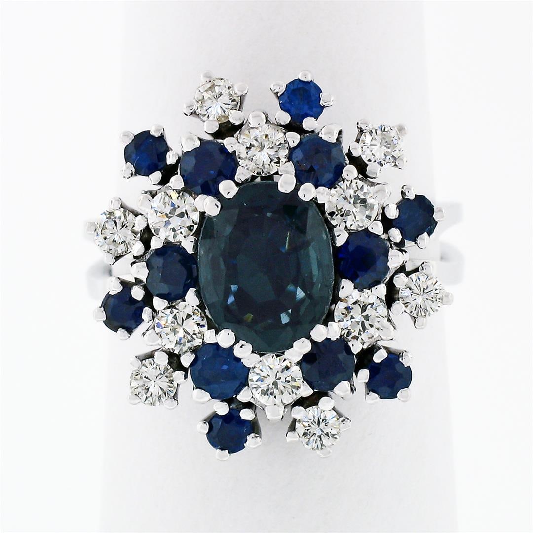 Vintage 18k White Gold 3.15 ctw Oval Sapphire Round Diamond Tiered Cluster Ring: A vintage 18k white gold cocktail ring with 1 oval brilliant cut natural sapphire (1.55ct) surrounded with 12 round cut natural sapphires (1.10ctw) and 12 round brilliant cut natural diamonds (F/G - V