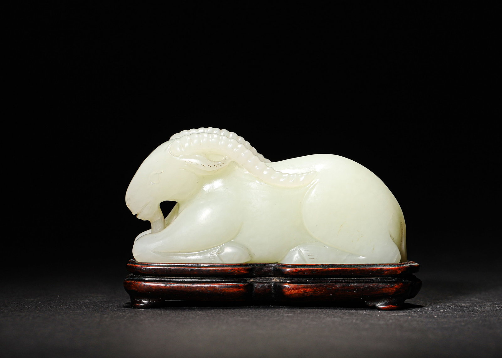 MID-QING DYNASTY, WHITE HETIAN JADE RECUMBENT RAM ORNAMENT: 3.38" x 1.62" (8.5cm x 4.0cm) | 0.33lbFinely carved from warm, creamy white Hetian jade, this recumbent ram features a plump, serene form and smooth, fluid carving, embodying the technical sophisticat