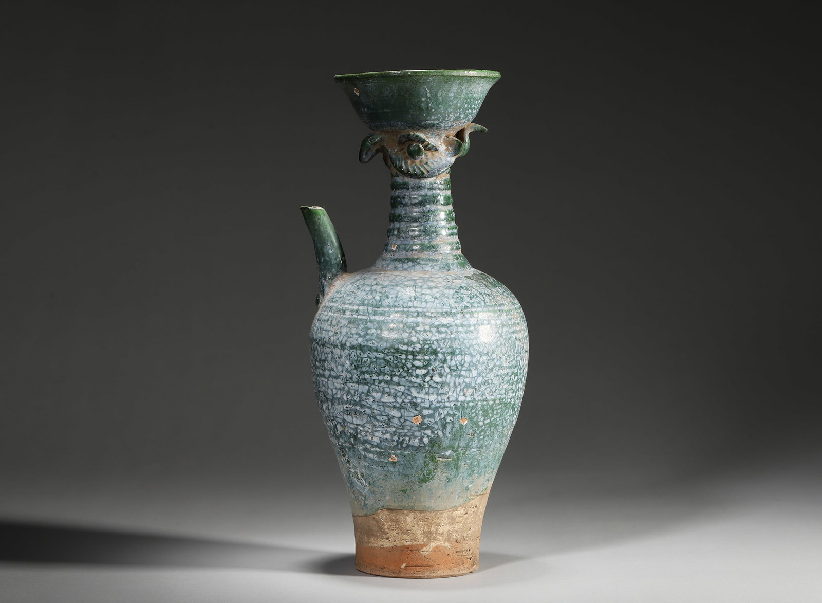 LIAO DYNASTY, GREEN-GLAZED EWER WITH PHOENIX-HEAD SPOUT AND ICE-CRACKLE PATTERNS: 5.12" x 12.6" inches (13.0 cm x 32.0 cm)This robust ewer features a weathered green glaze with stunning ice-crackle patterns, crowned by a vivid phoenix-head spout. A perfect blend of Liao nomadic art