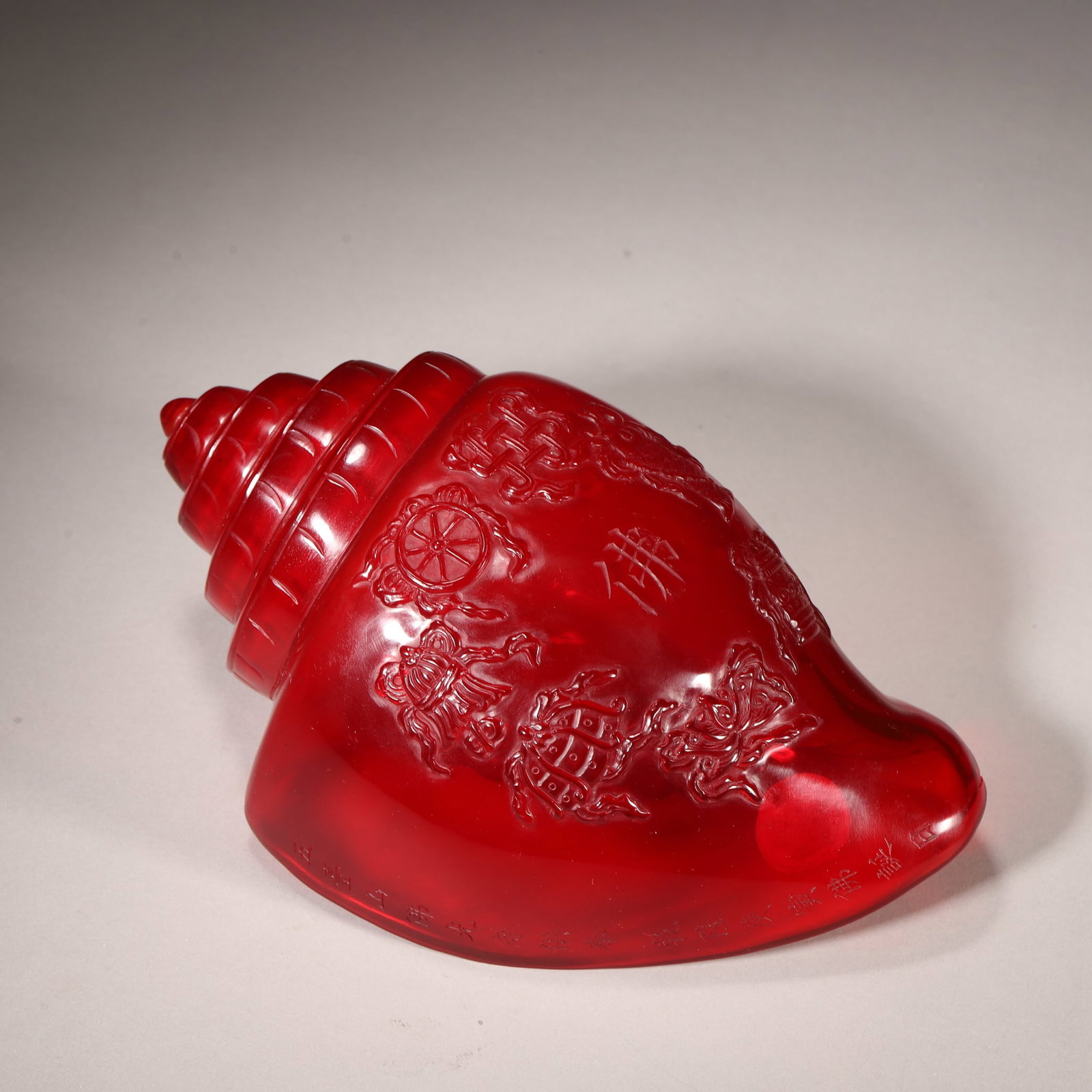 A Red Glass Conch with Eight Auspicious Symbols, Qianlong Period, Qing Dynasty: L 21.0cm (8.27 in); W 11.0cm (4.33 in); H 9.0cm (3.54 in); Wt 809.0g (1.78 lb)This conch is carved from a whole piece of red glass, with a vivid, gem-like transparency. The exterior features raised Ei