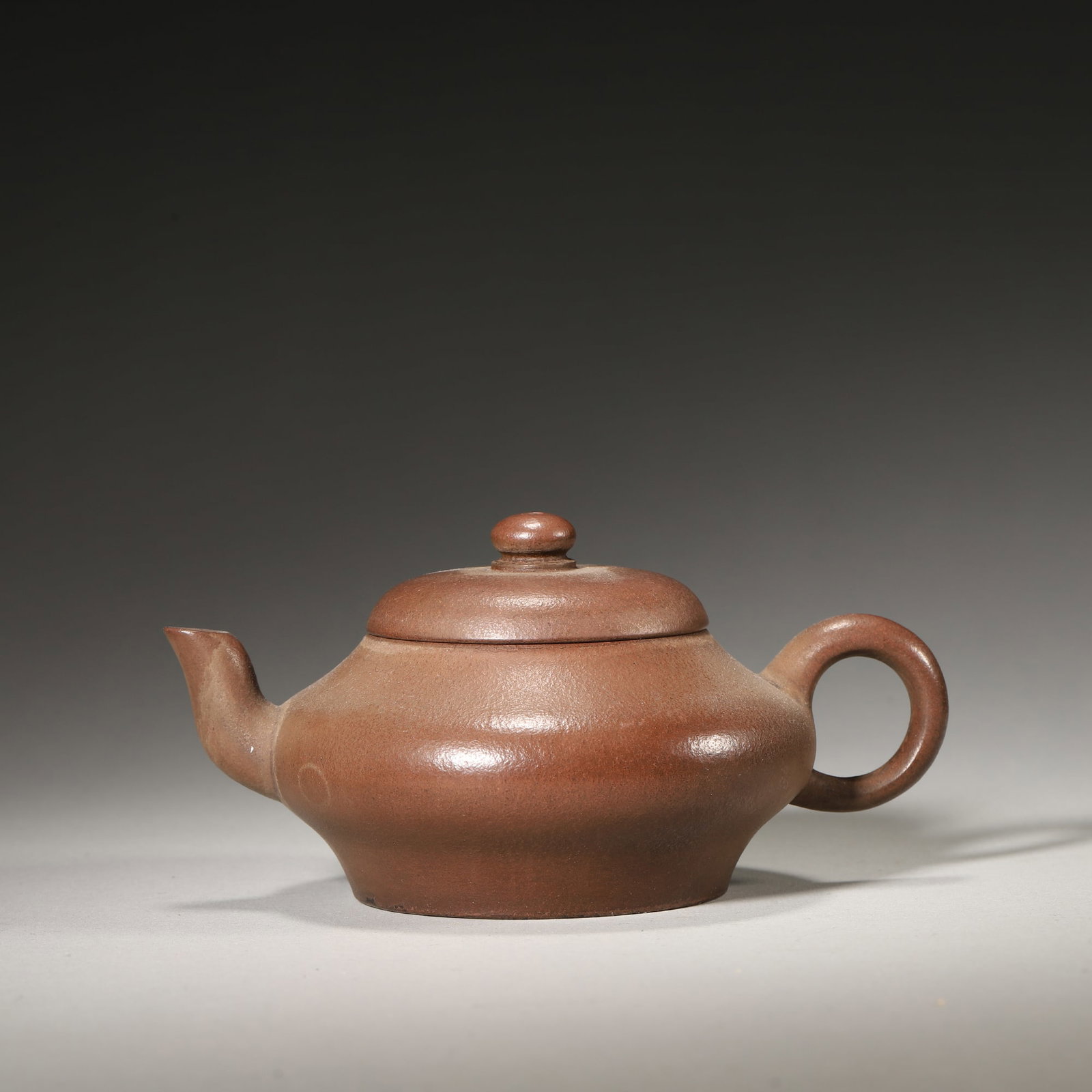 A Zisha Flat Teapot, Qing Dynasty: L 12.5cm (4.92 in); H 6.0cm (2.36 in)This teapot is crafted from fine Zisha clay in a simple flat form, with a calligraphy mark at the base. It is an elegant example of Qing Dynasty scholar tea ware w