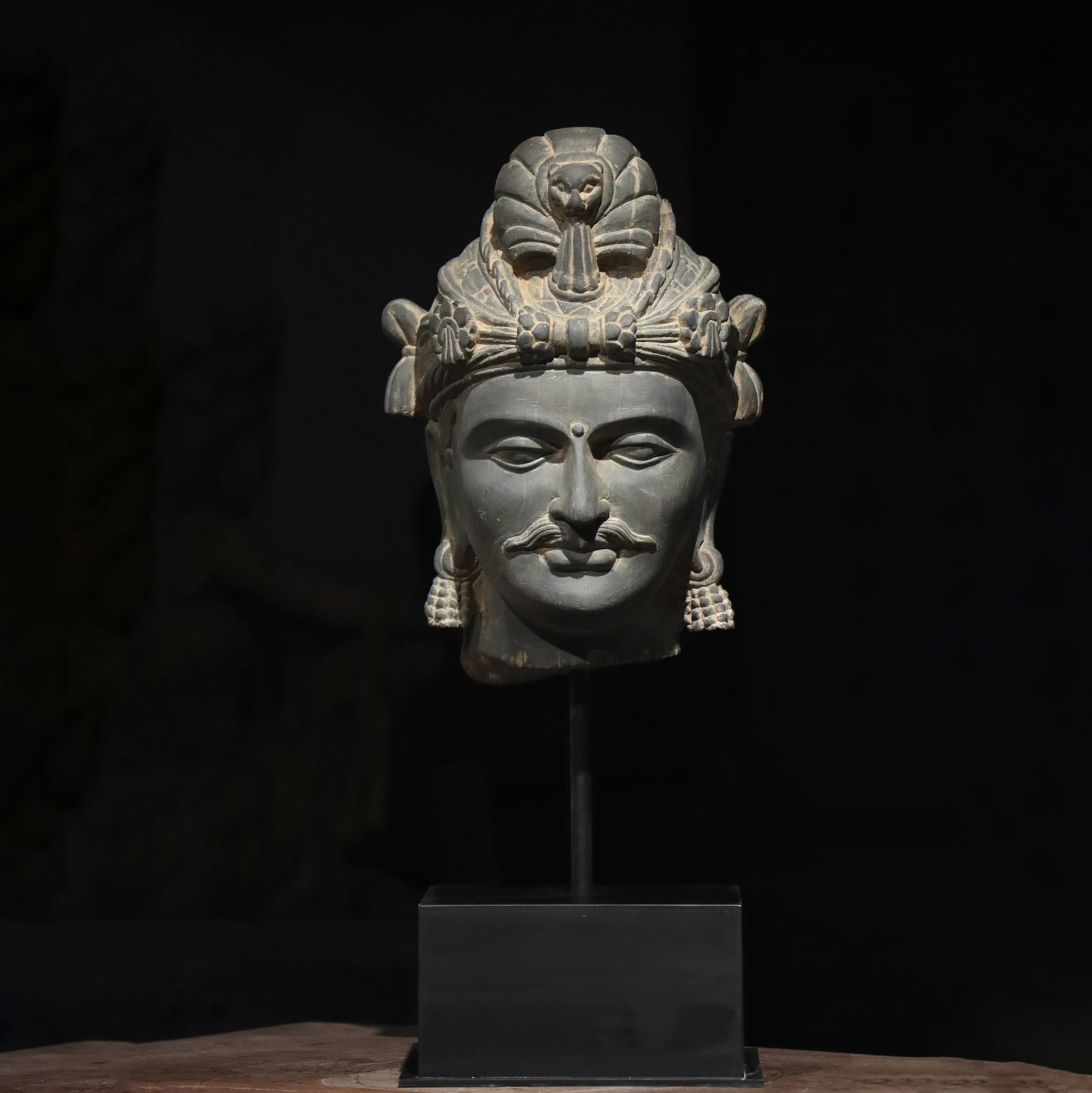 A Grey Schist Head of a Bodhisattva, Gandhara, 2nd-3rd Century: H (with base) 53cm (20.87 in); H (net) 32cm (12.6 in)A grey schist head of a bodhisattva from Gandhara, 2nd-3rd century. Carved from fine grey schist, it features a richly adorned crown with a fan-sha