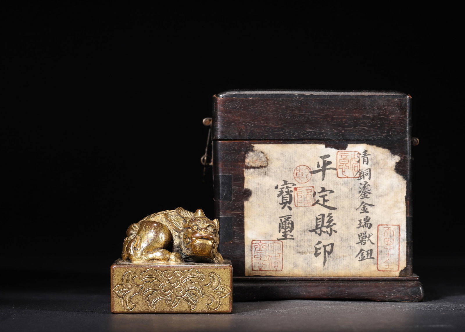 QING DYNASTY, A GILT-BRONZE SEAL WITH MYTHICAL BEAST KNOB, INSCRIBED 'PING DING XIAN YIN', FITTED: 2.05" x 2.05" x 1.97" (5.2cm x 5.2cm x 5.0cm) 1.422lb (645g)This gilt-bronze official seal features a recumbent mythical beast knob and nine-fold seal script. It retains its original hardwood storage 