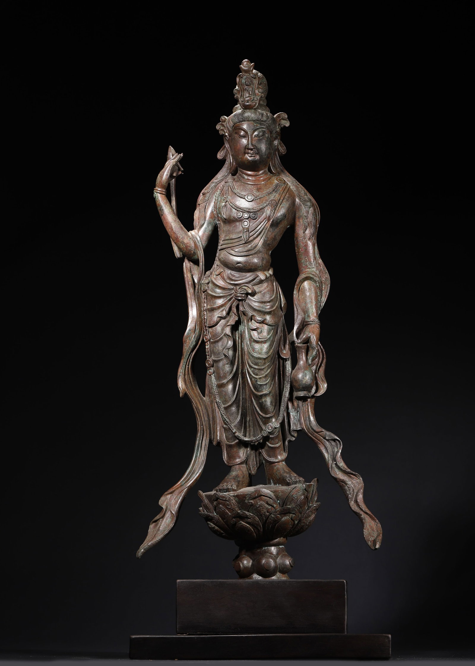TANG DYNASTY BRONZE STANDING STATUE OF GUANYIN BODHISATTVA: 22.44in (57.0cm)This Tang Dynasty bronze Guanyin stands in a graceful S-curve, embodying the Tang aesthetic of plump elegance. Adorned with a crown, necklaces, and flowing robes, it holds a pure vase,
