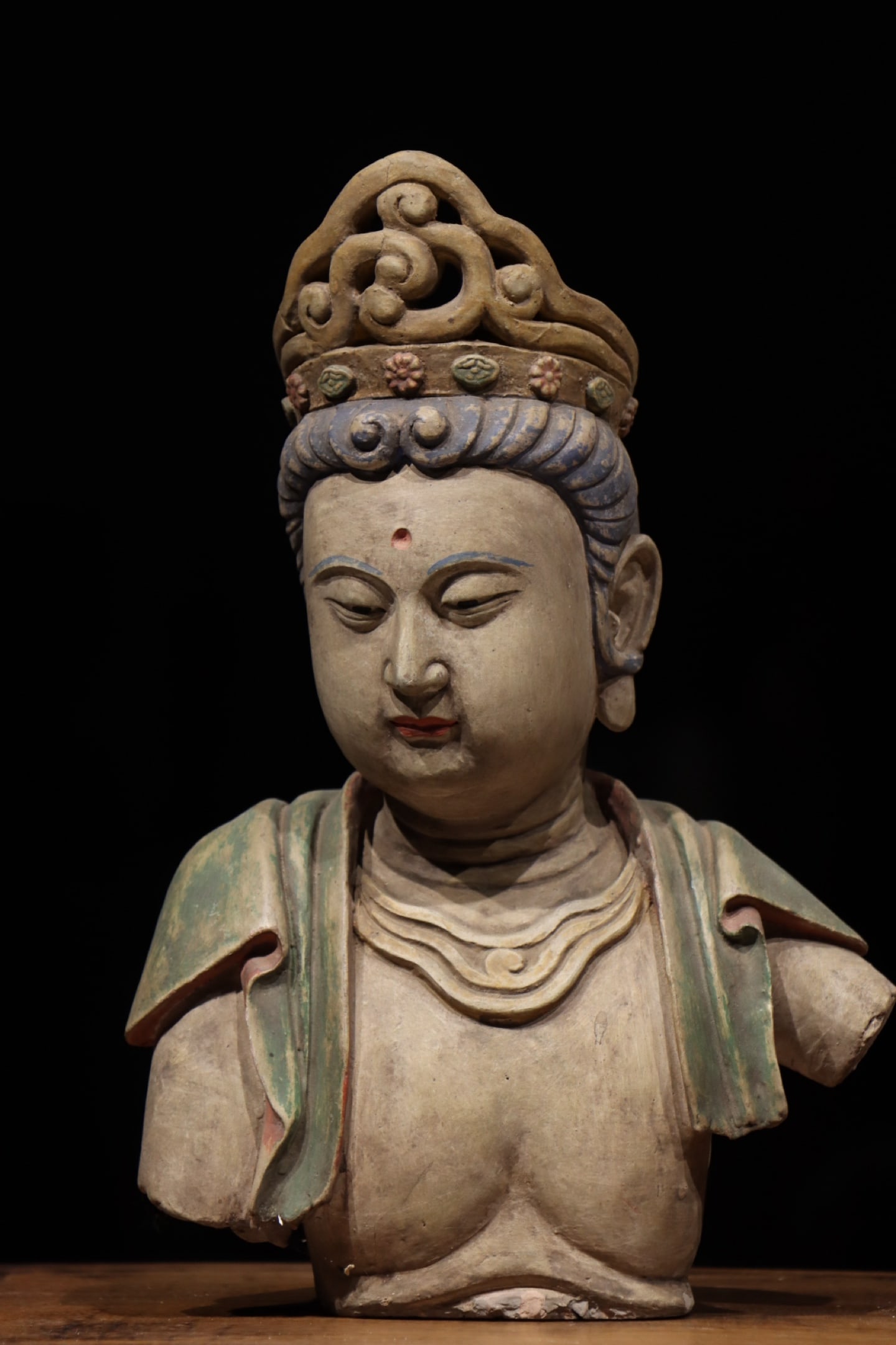 A PAINTED CLAY BUST OF A BODHISATTVA, MING DYNASTY, 14TH–15TH CENTURY: Height: 14.17in (36.0cm)Carved from clay and painted, this Ming Bodhisattva bust features a serene plump face, an openwork crown, and vibrant mineral pigments. Its flowing drapery embodies the elegant