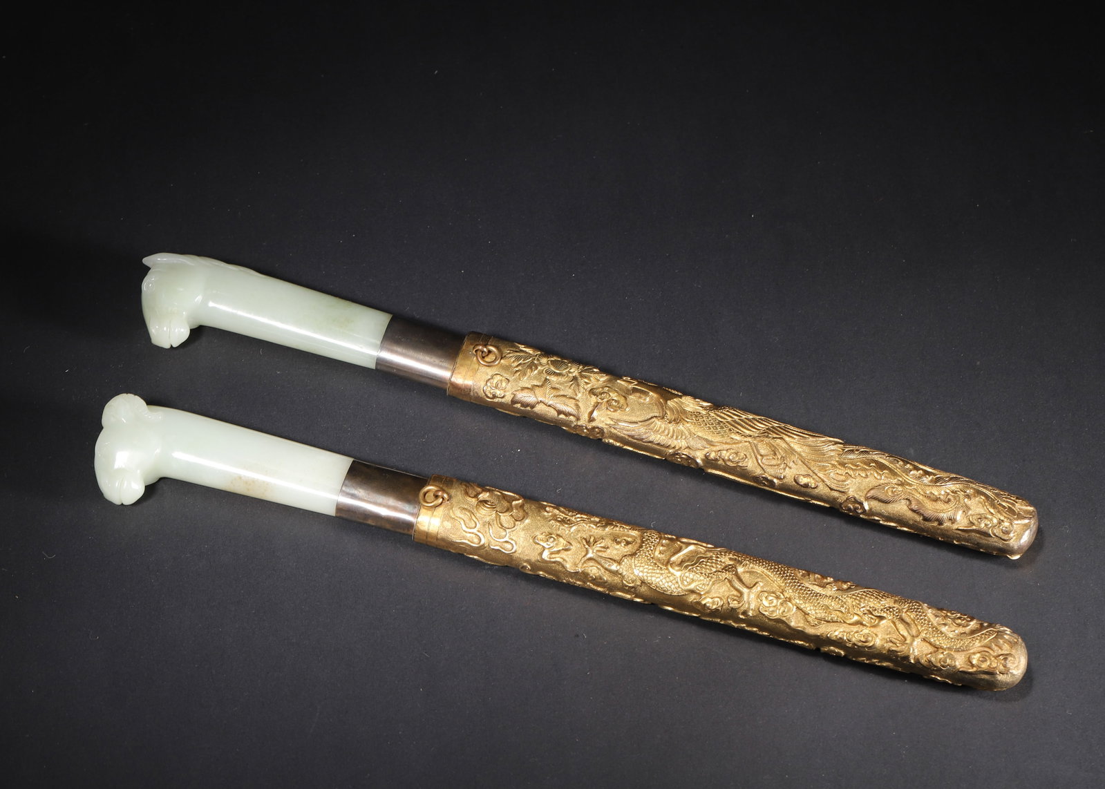 QING DYNASTY PAIR OF PALE GREEN JADE-HANDLED KNIVES WITH GILT-BRONZE DRAGON AND PHOENIX SCABBARDS: 12.60in x 1.38in (32.0cm x 3.5cm) 1.217lbs (552g)A fine pair of Qing Dynasty pale green jade-handled knives, with horse and ram head finials, housed in gilt-bronze scabbards carved with dragon and pho